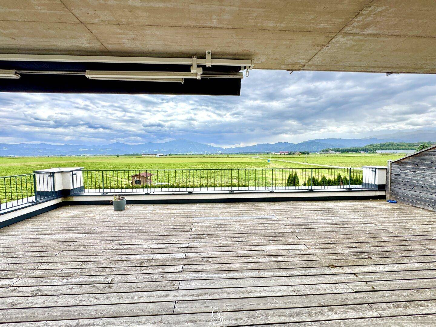 Große Holzterrasse mit Metallgeländer und weitem Blick über Felder und die Bergkette.