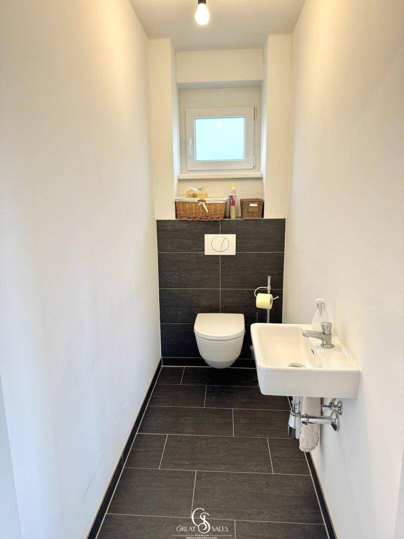 Gäste-WC mit wandhängender Toilette, kleinem Waschbecken und einem Fenster für Tageslicht.