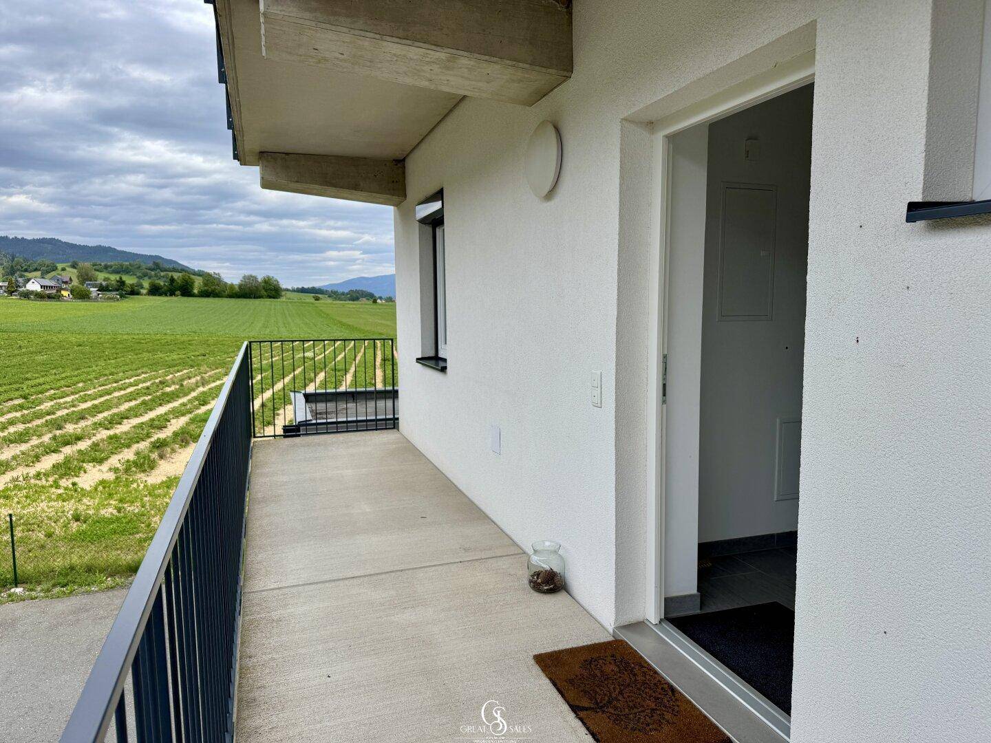 Kleiner Betonbalkon mit Geländer und Zugang zur Wohnung, umgeben von grünen Feldern.