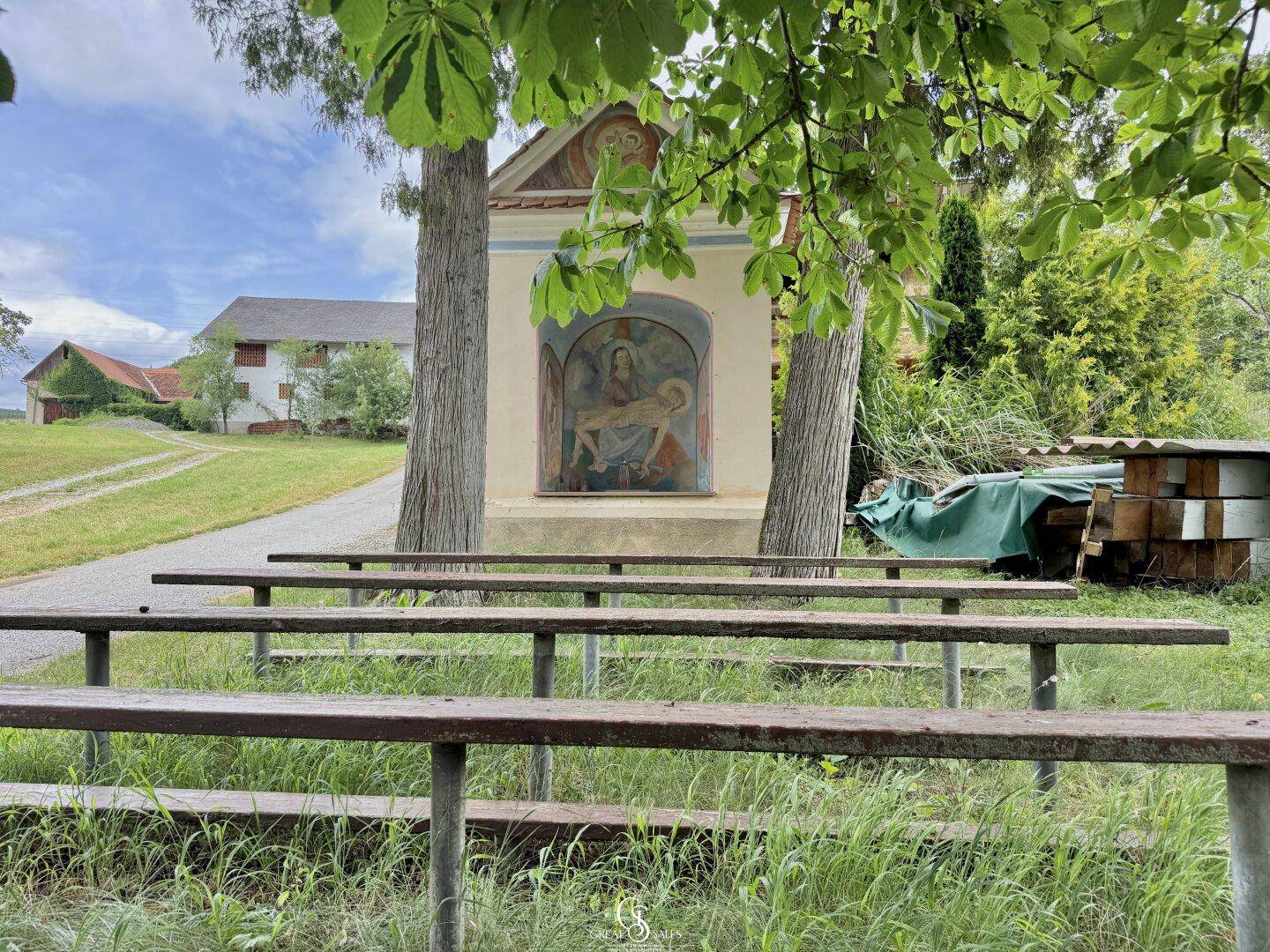 Kleine Kapelle mit Bänken unter Bäumen in einer ländlichen Umgebung.