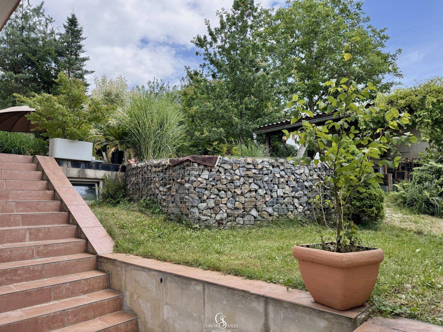 Gartenbereich mit Steintreppe, Gabionenwand und Topfpflanzen.