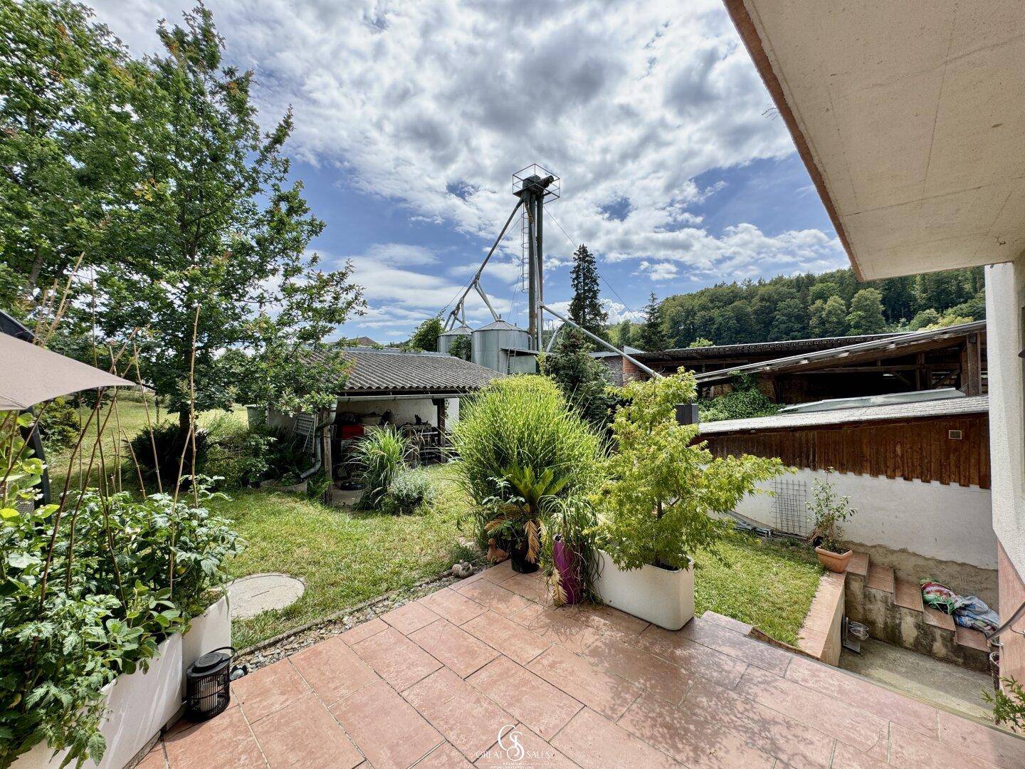 Gepflasterte Terrasse mit Pflanzen und Blick auf landwirtschaftliche Silos und Gebäude.