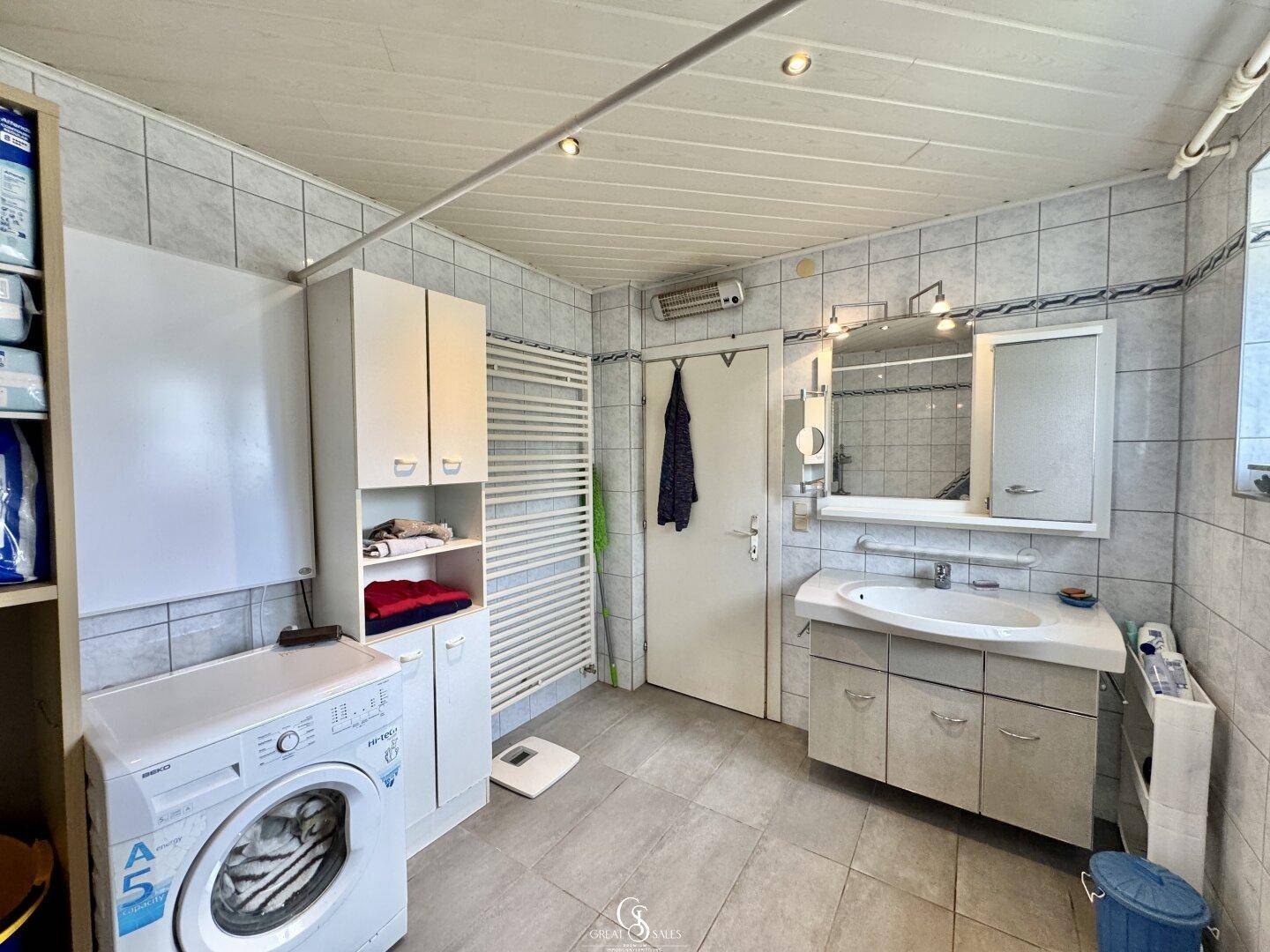 Geräumiges Badezimmer mit Fliesenboden, Waschmaschine, Heizkörper und Spiegelschrank.
