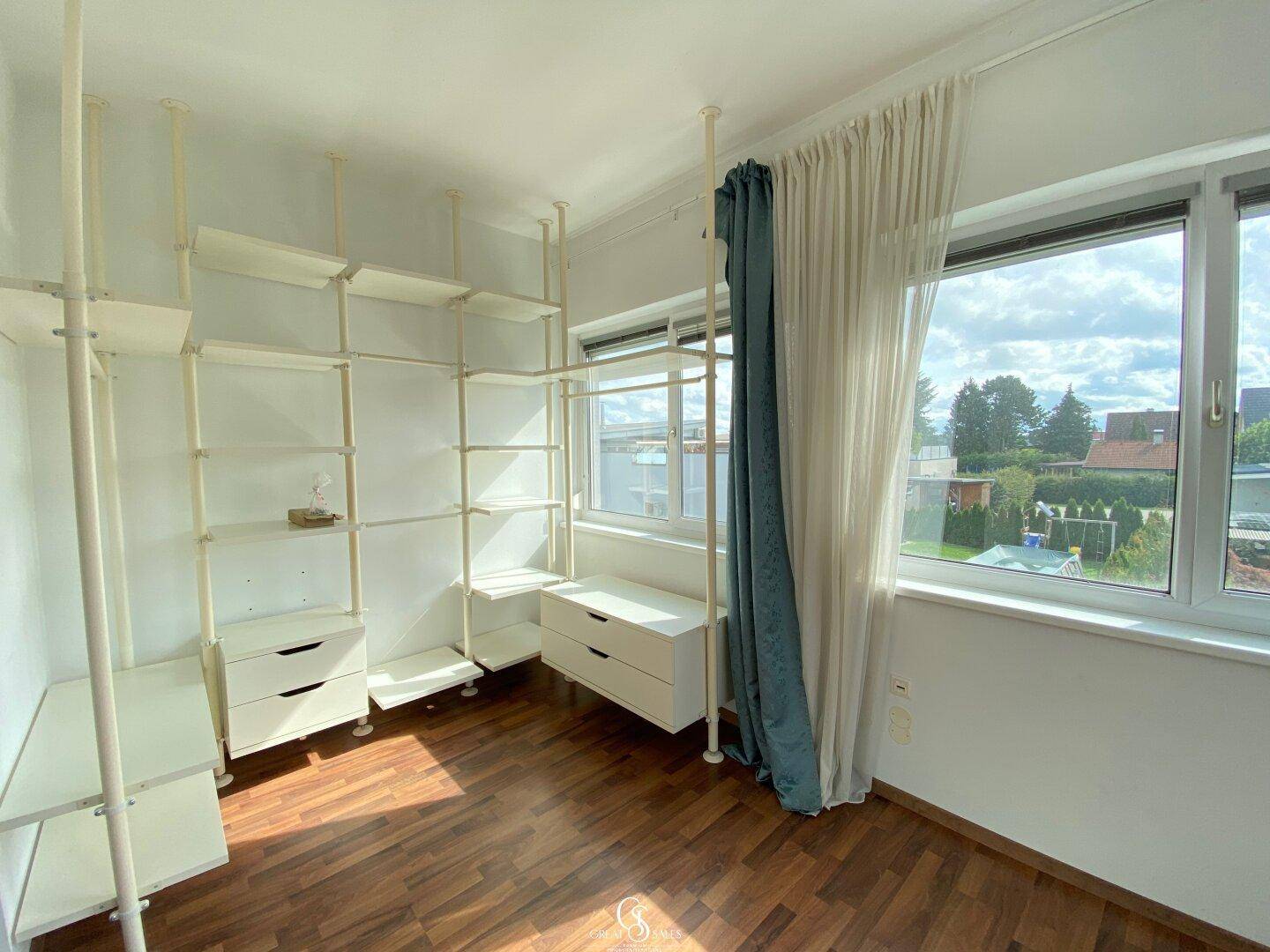 Helles Zimmer mit Regalsystem, Holzboden und großen Fenstern mit Blick ins Grüne.