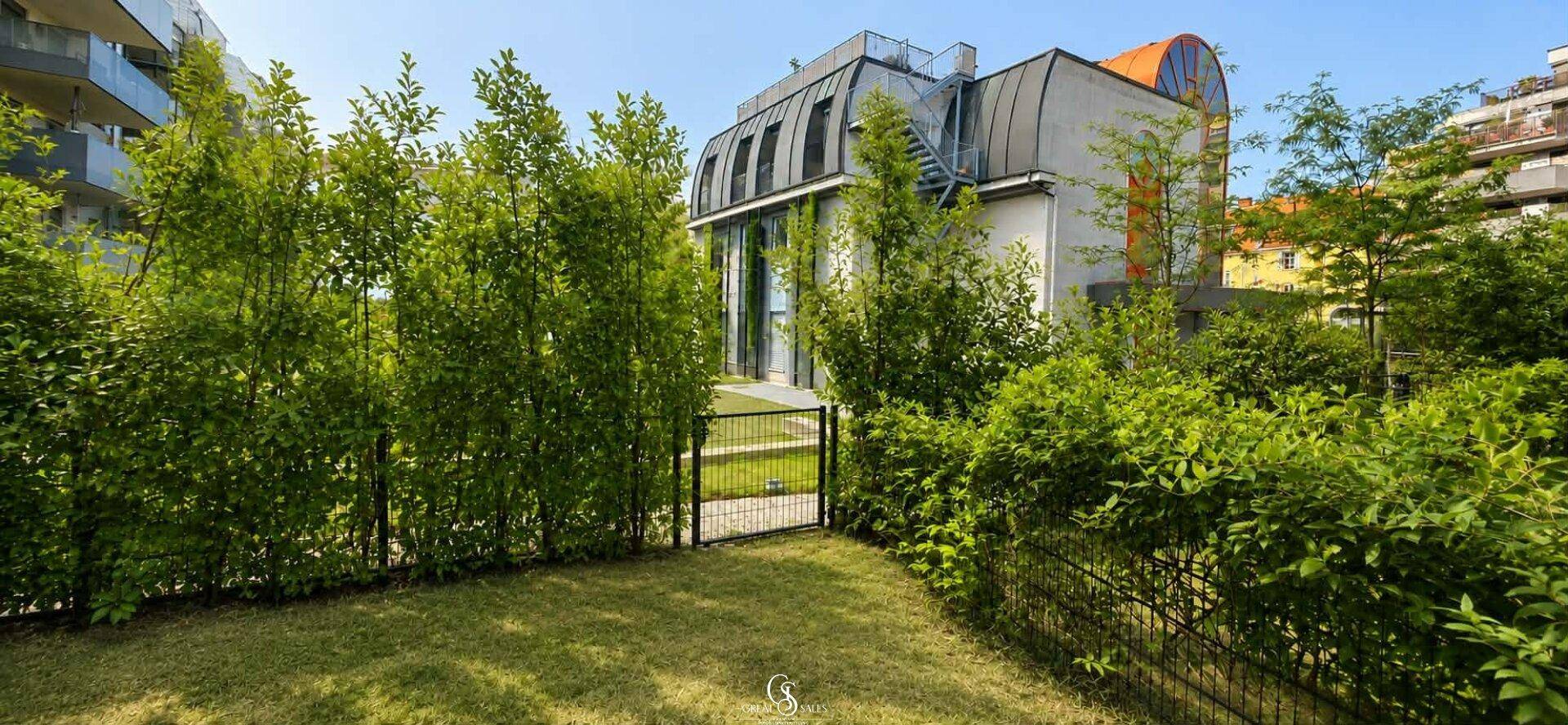 Modernes Gebäude mit grüner Bepflanzung und gepflegter Außenanlage unter blauem Himmel.