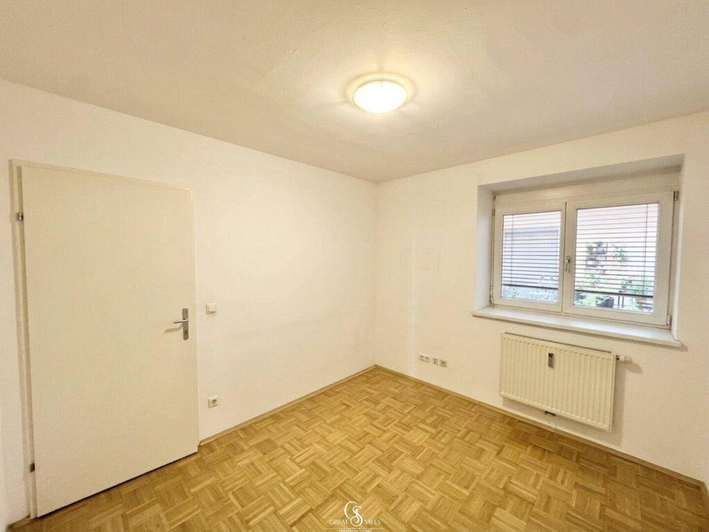 Helles Schlafzimmer mit Parkettboden und Fenster, das den Blick ins Grüne freigibt.