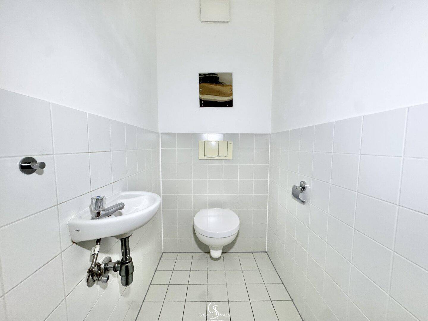 Separates WC mit wandhängender Toilette, kleinem Waschbecken und weißen Fliesen.