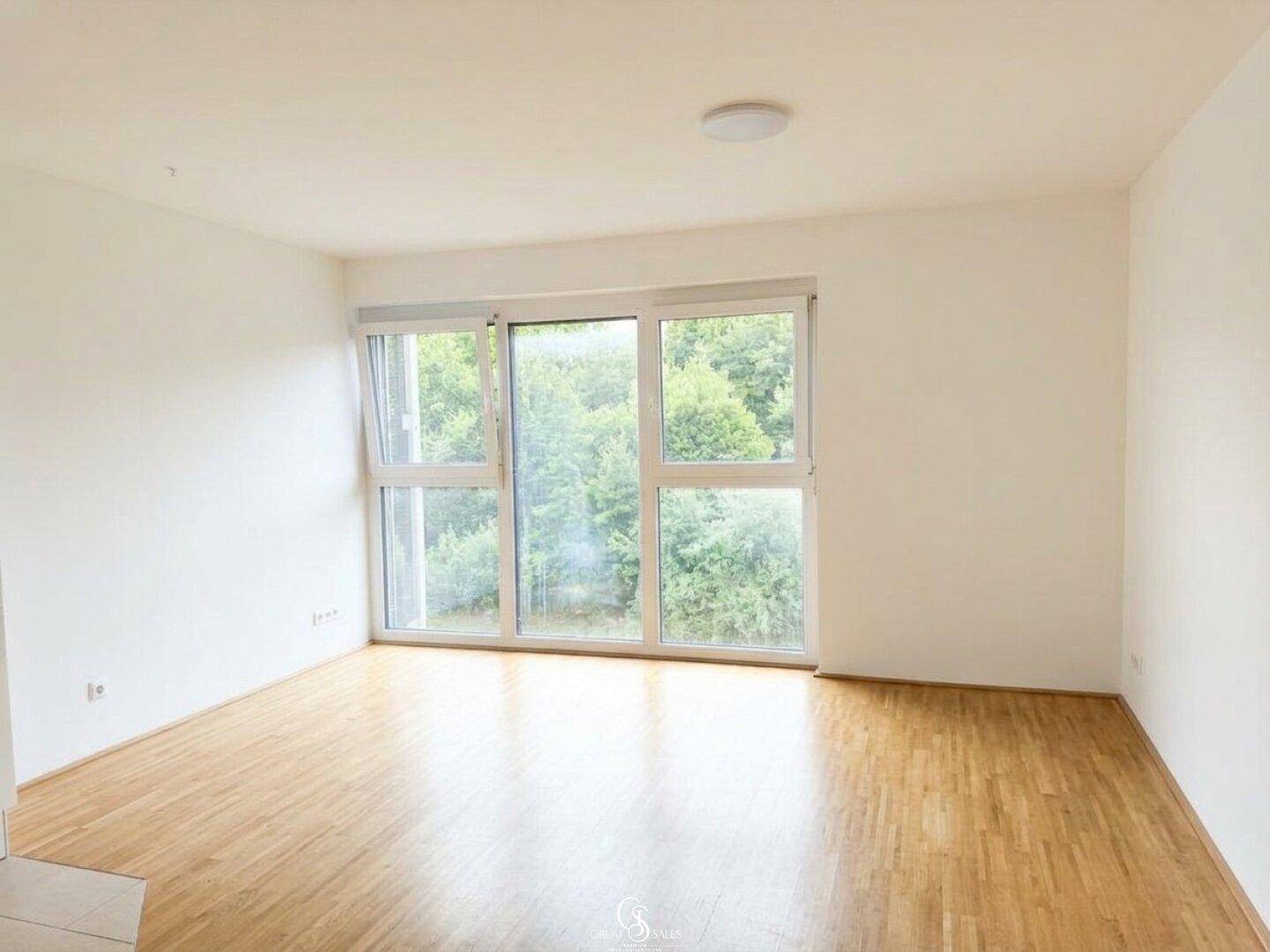 Lichtdurchflutetes Wohnzimmer mit Holzboden und großen Fenstern, die einen schönen Blick ins Grüne bieten.
