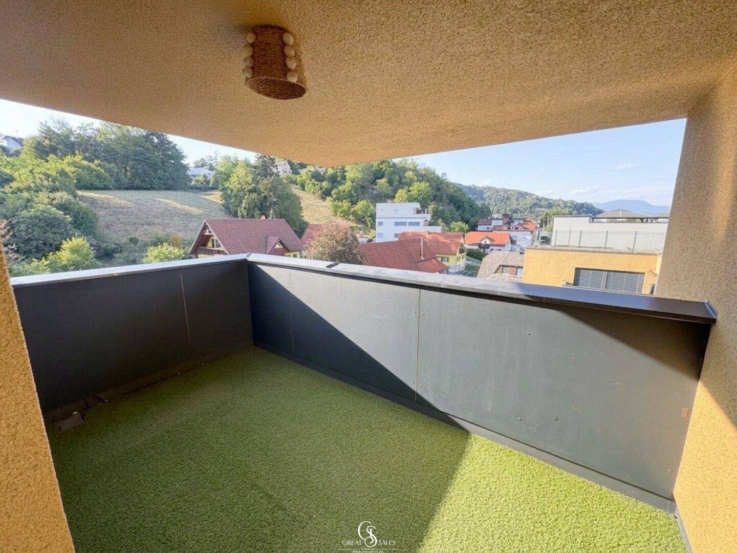 Balkon mit Kunstrasen und Blick auf die umliegende Hügellandschaft und die Dächer der Nachbarhäuser.