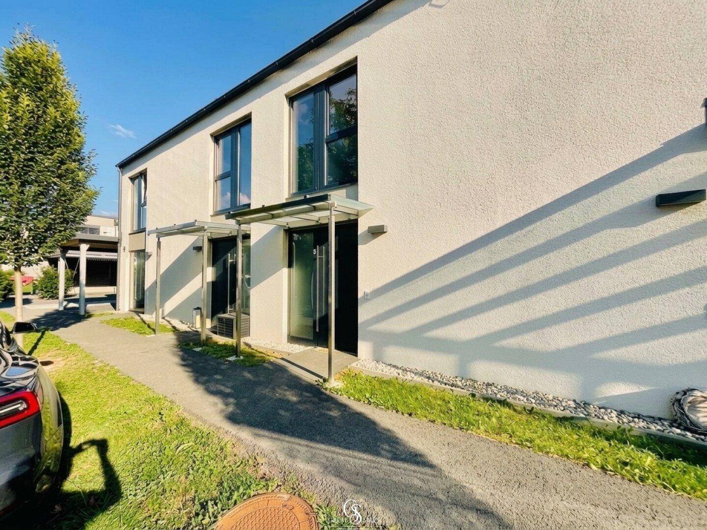 Moderne Reihenhausfassade mit großen Fenstern und Vordächern, umgeben von Grünflächen.