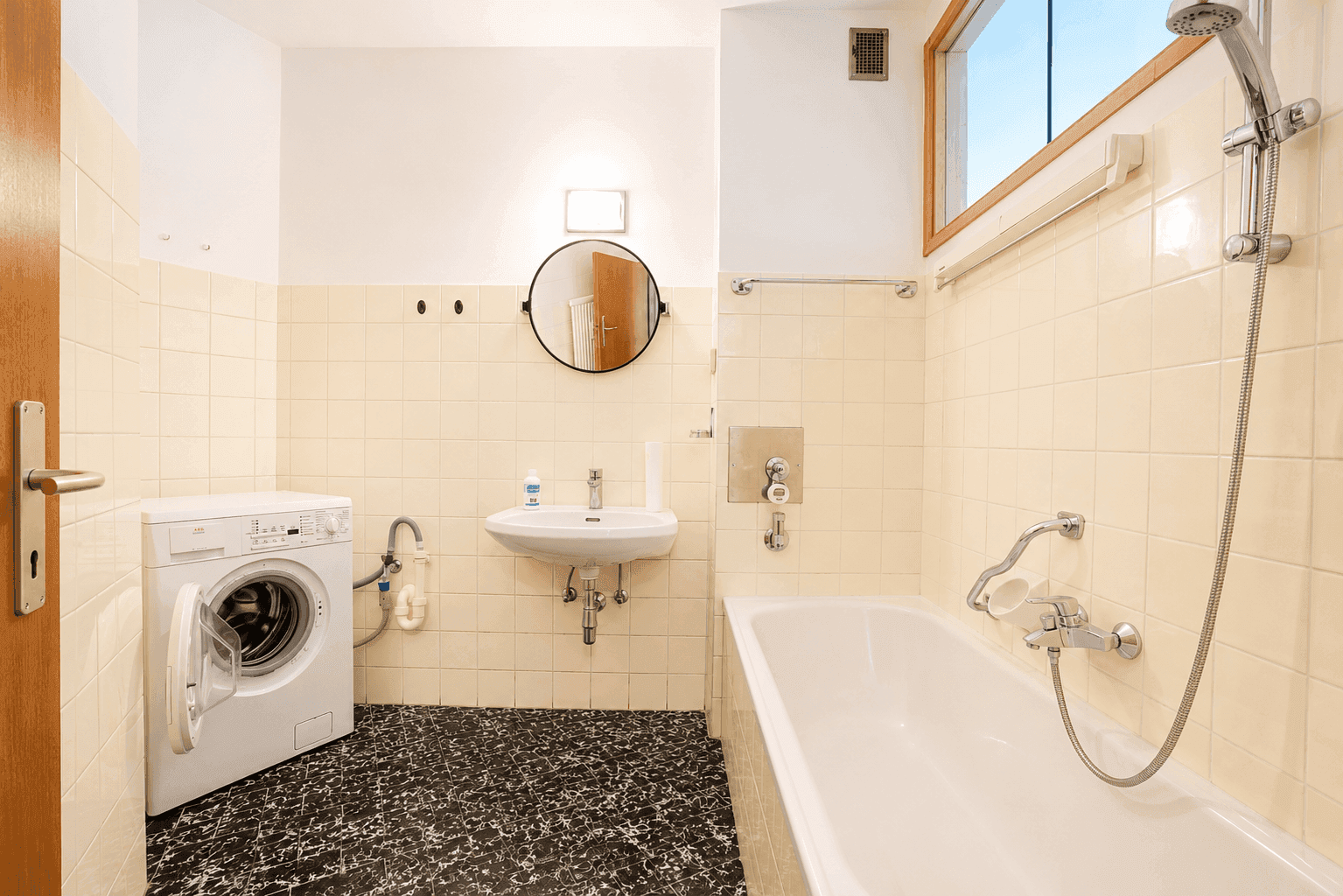Badezimmer mit Badewanne, Waschbecken, Waschmaschine und Fliesen in Beige und Schwarz im 80er-Jahre-Stil.