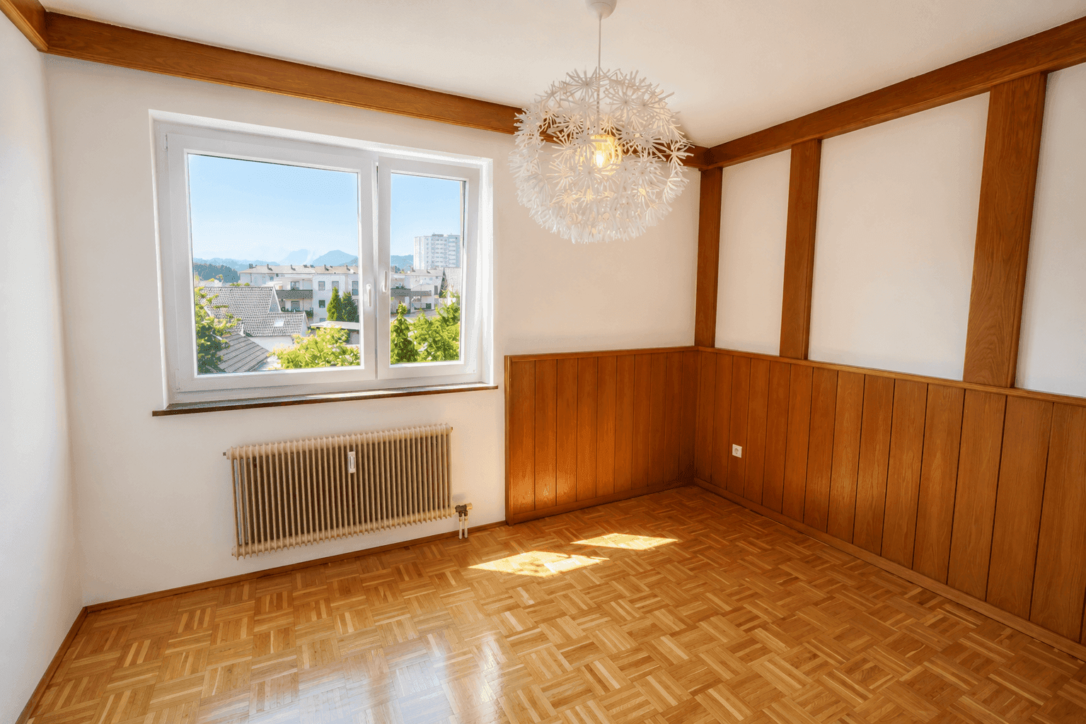 Freundliches Zimmer mit Parkettboden, Holzvertäfelung und einem weiten Blick ins Grüne und auf die Berge.