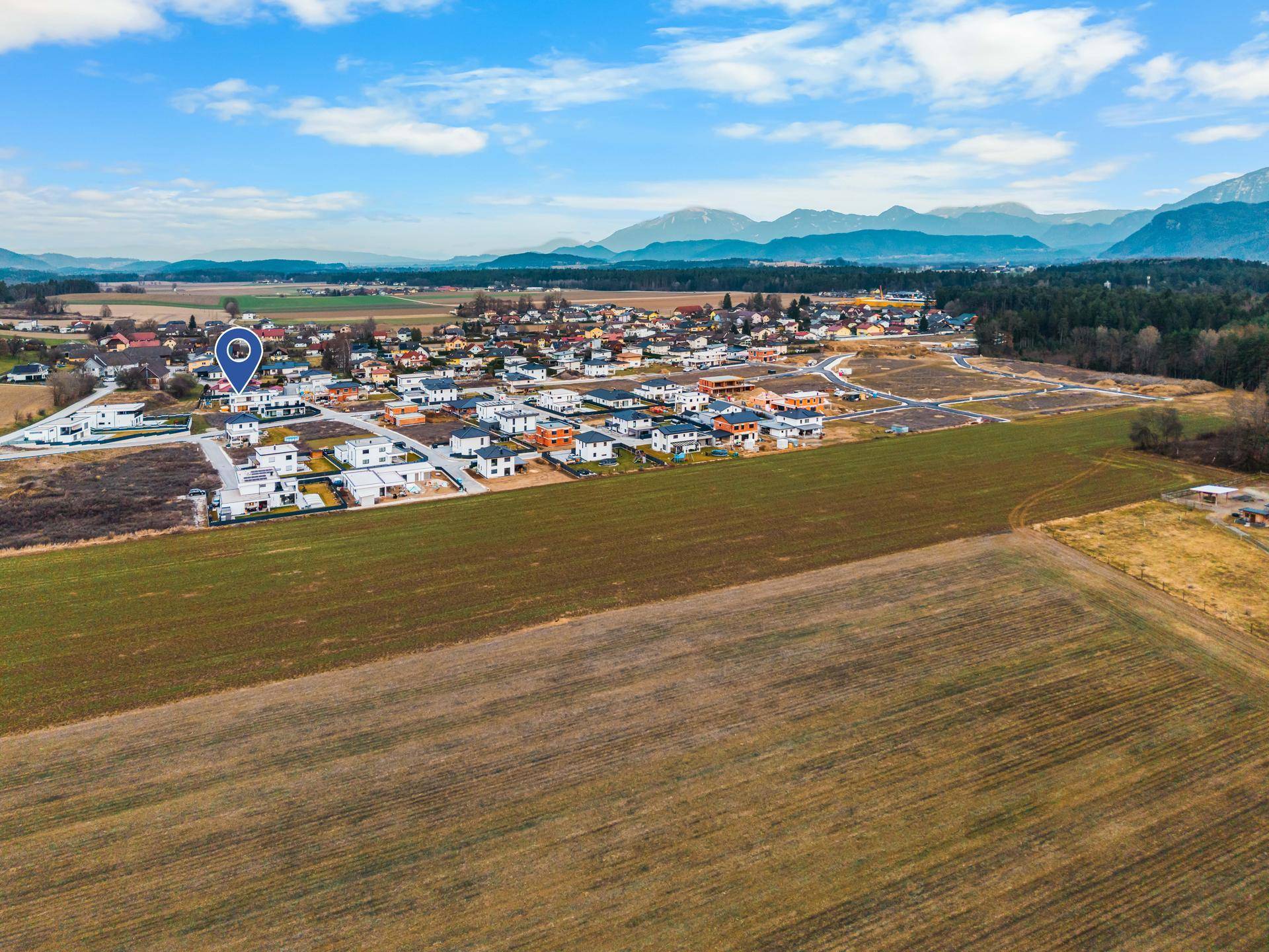 Luftaufnahme einer Wohnsiedlung mit Neubauten, umgeben von Feldern und Bergen im Hintergrund.
