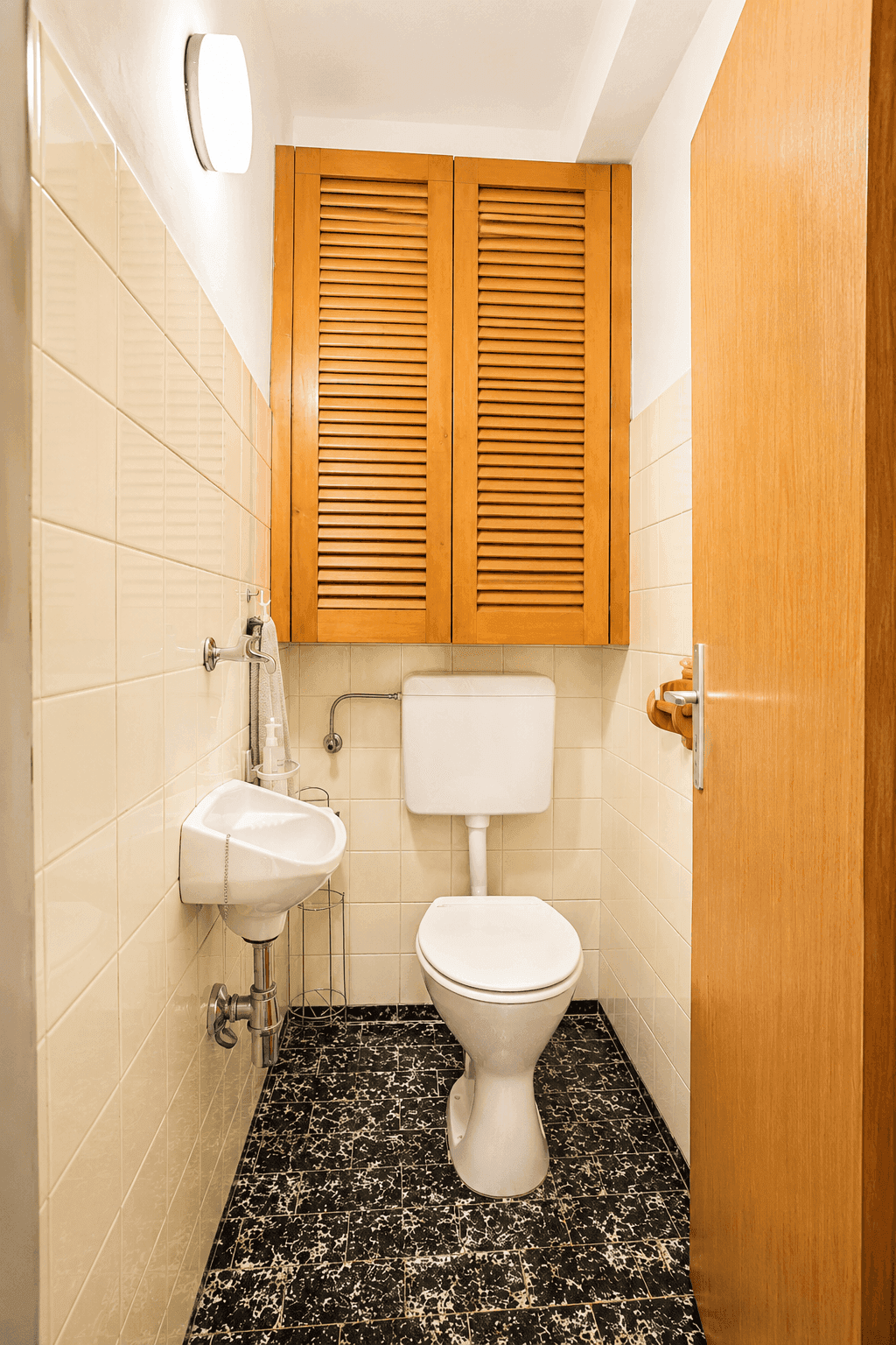 Kleines Gäste-WC mit WC, Waschbecken und Fliesen in hellem Beige und Schwarz im 80er-Jahre-Stil.