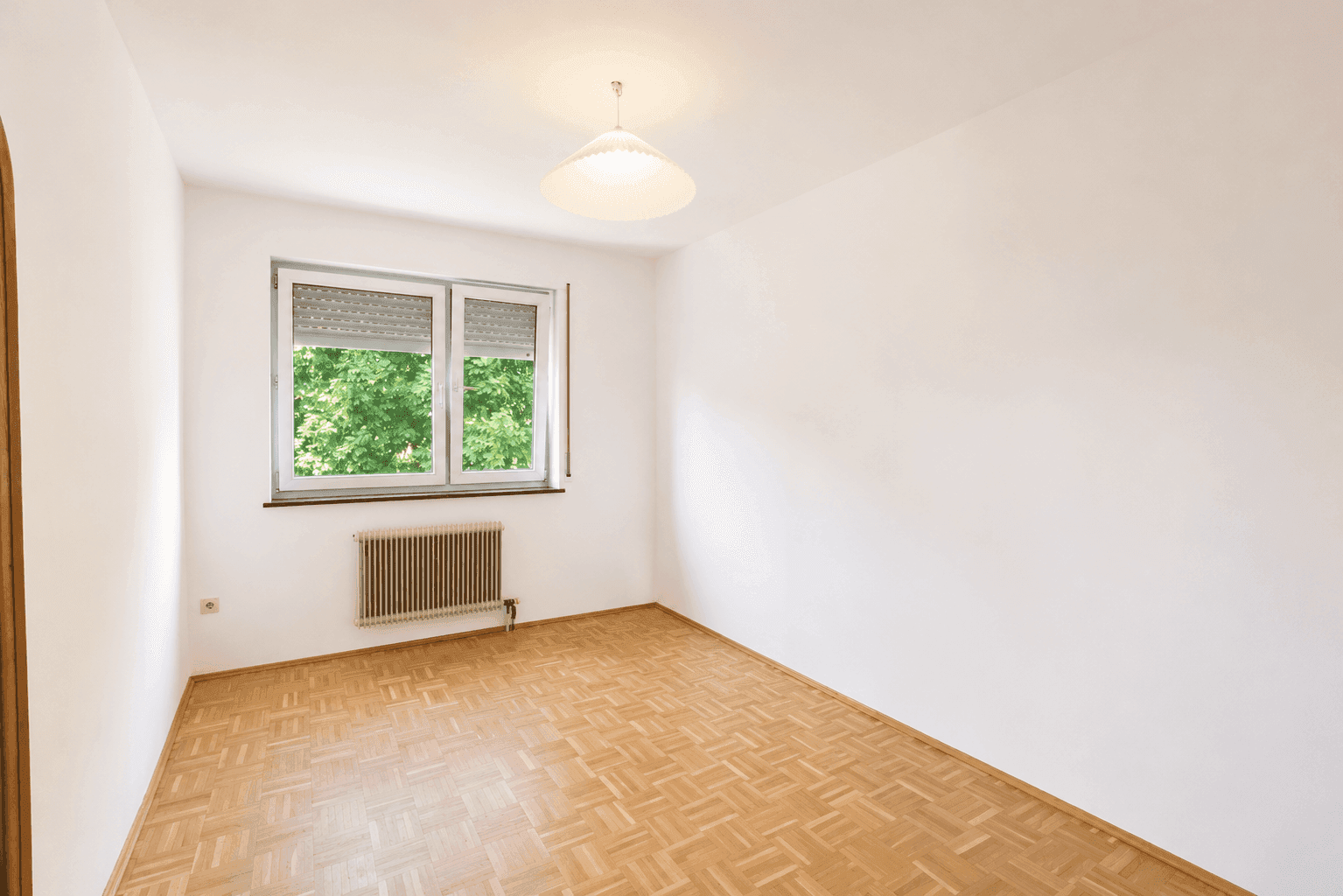 Leeres Schlafzimmer mit Parkettboden, Fenster mit Rollladen und einem beruhigenden Blick ins Grüne.