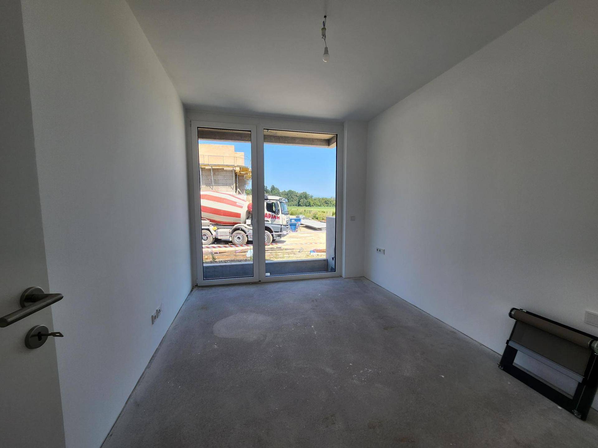 Unfertiger Raum mit Betonboden und großer Fenster-Schiebetür zum Außenbereich.