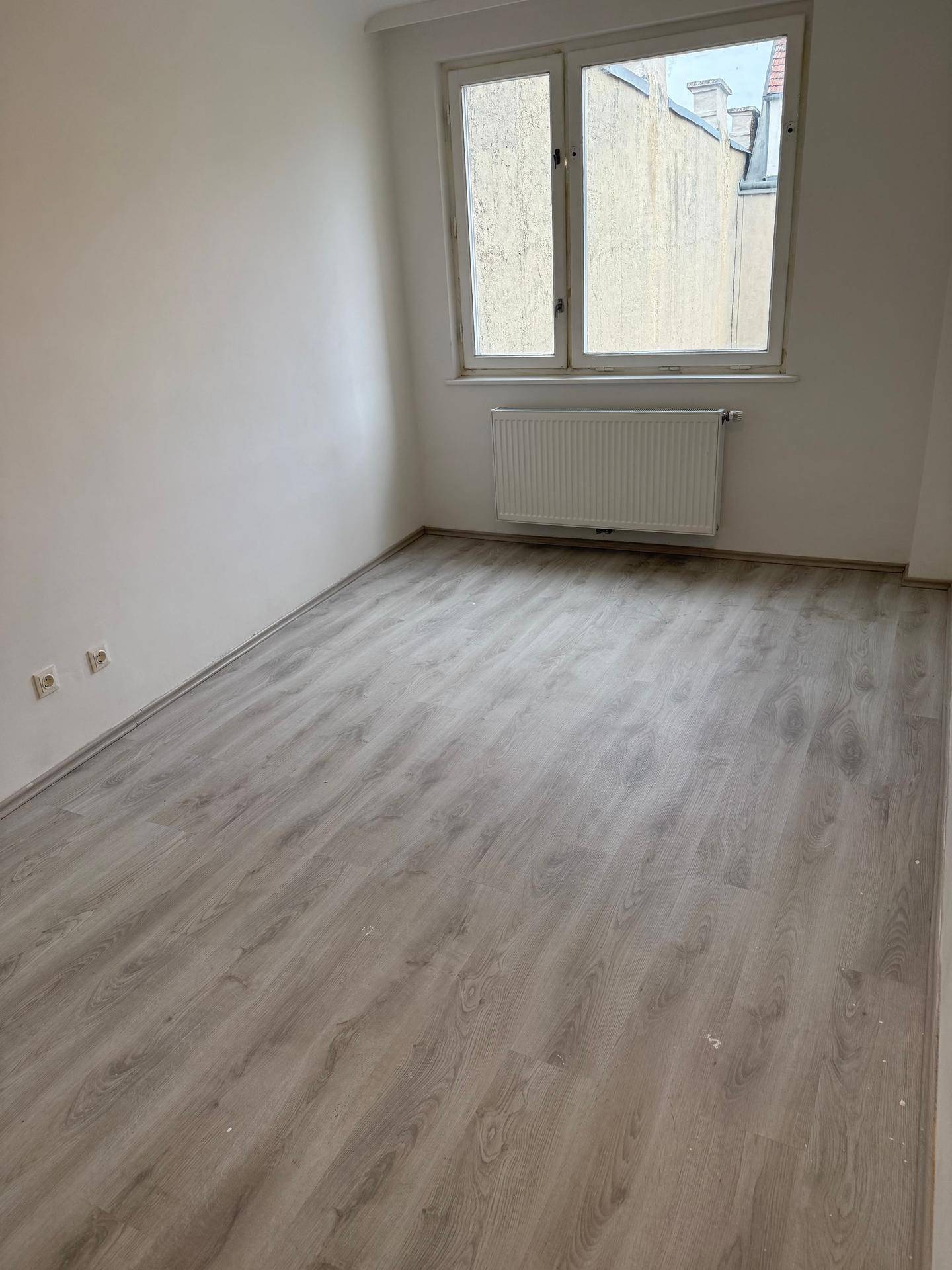 Unmöbliertes Zimmer mit hellem Laminatboden und Fenster, das Tageslicht hereinlässt.