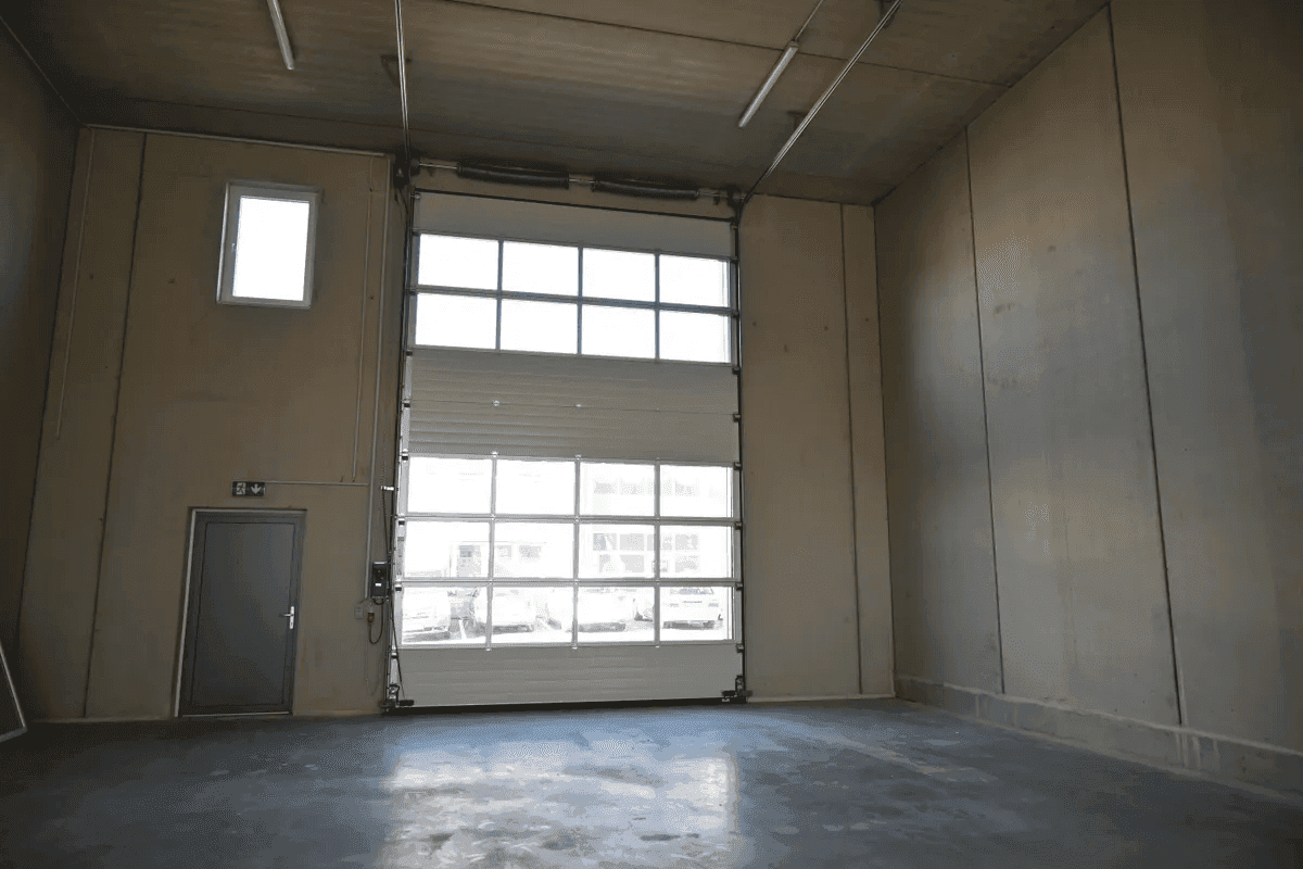 Leere Industriehalle mit hohen Decken, Betonwänden und einem großen Sektionaltor mit Fenstern.