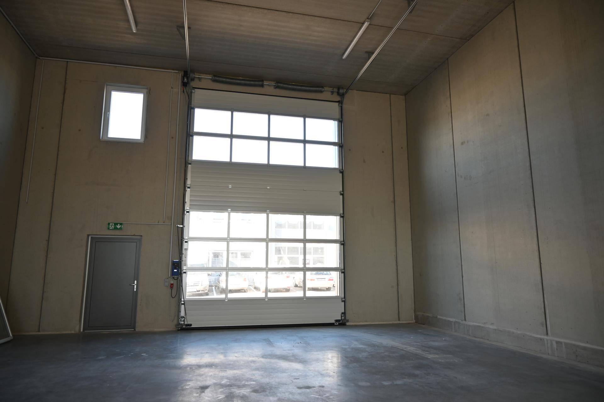 Geräumige Lagerhalle mit Betonwänden, großem Sektionaltor und viel Tageslicht durch die Fenster.