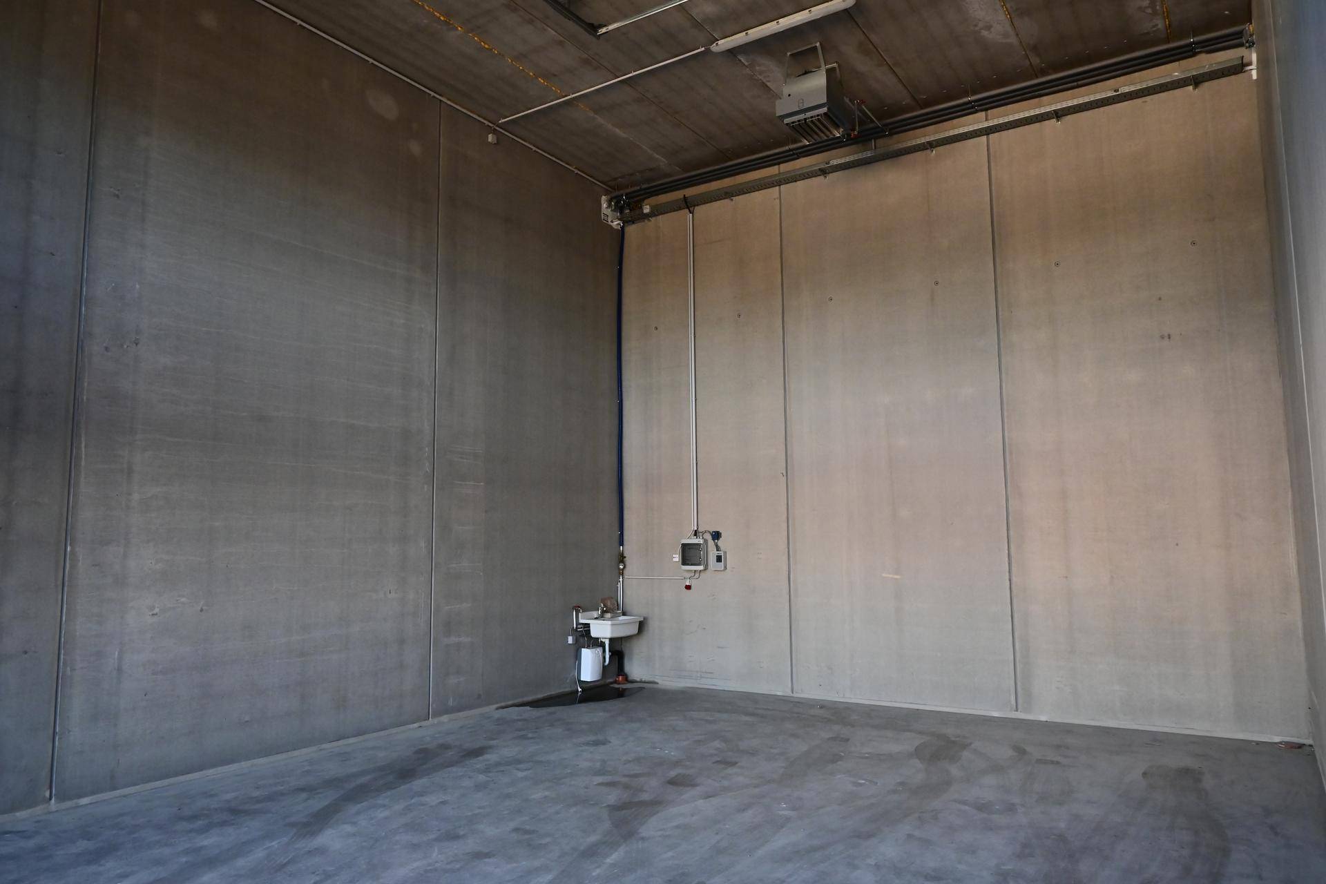 Leerer Industrieraum mit hohen Betonwänden, einem Waschbecken und sichtbaren Installationen an der Decke.