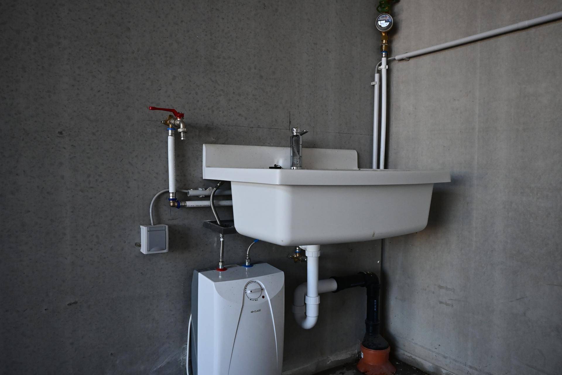 Nahaufnahme eines einfachen Waschbeckens mit Warmwasserbereiter und sichtbaren Wasserleitungen in einem Nutzraum.
