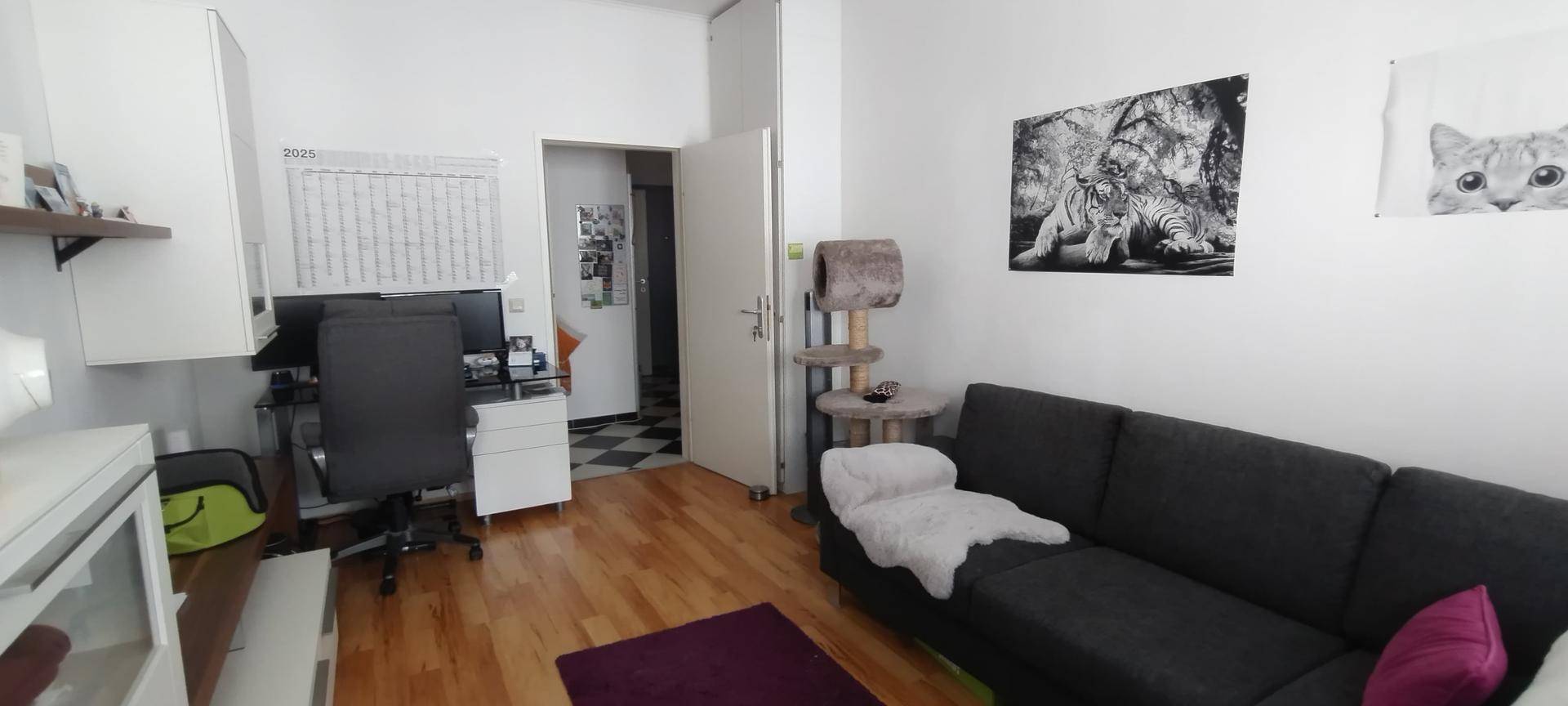 Wohnzimmer mit einem dunklen Sofa, einem Arbeitsbereich und einem Katzenbaum, hell und funktional eingerichtet.