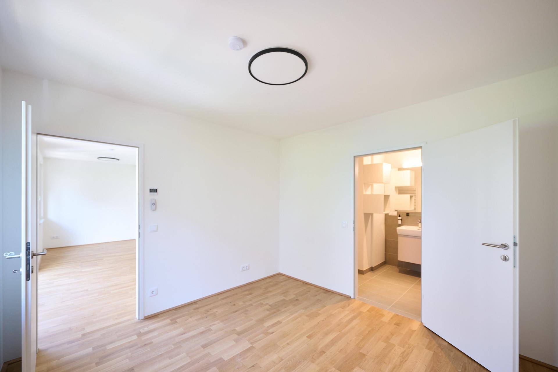 Helles, leeres Zimmer mit Parkettboden und Zugang zu einem weiteren Raum und Badezimmer.