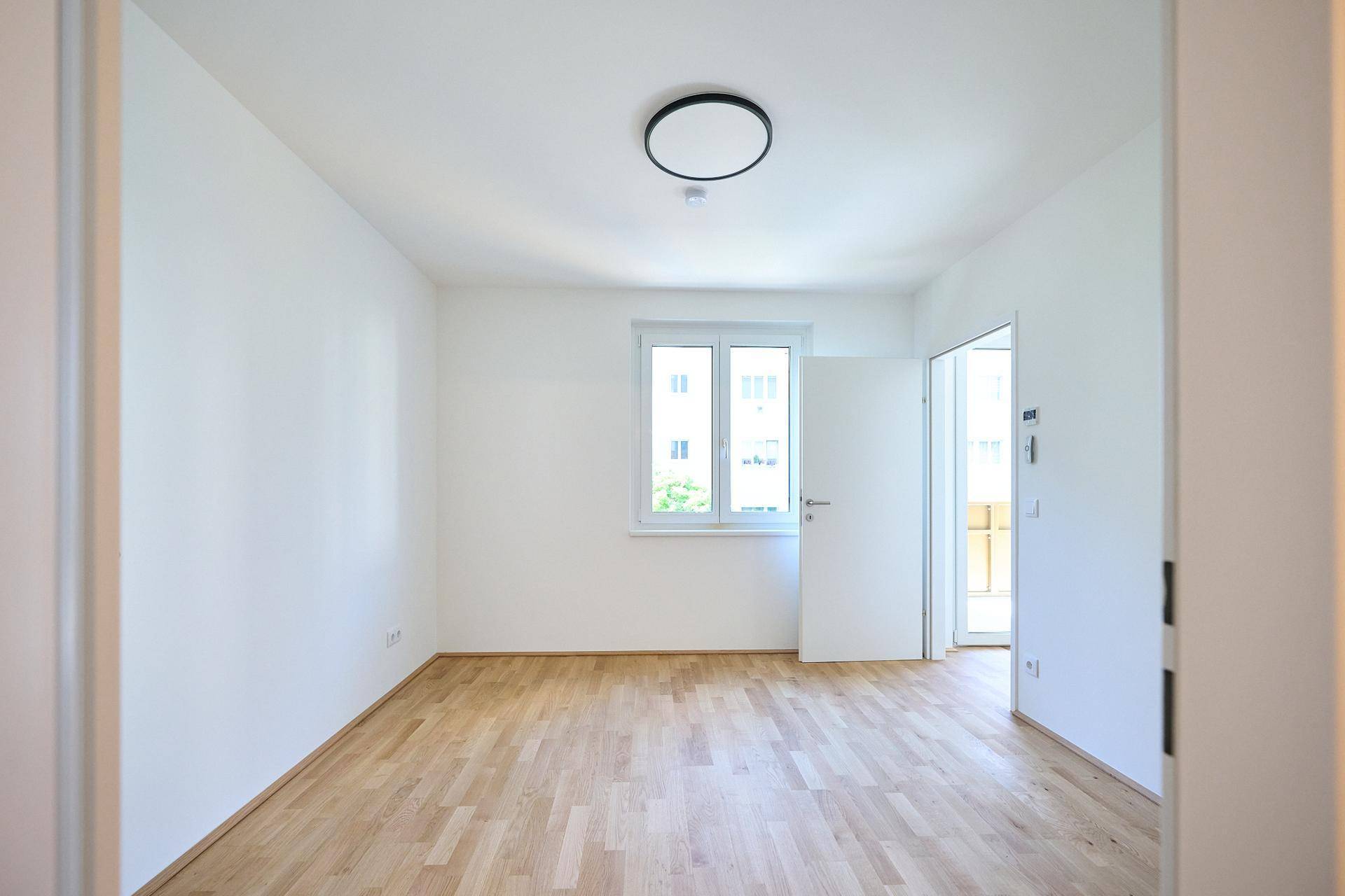Helles, leeres Zimmer mit Parkettboden, weißen Wänden und Zugang zu einem Balkon.