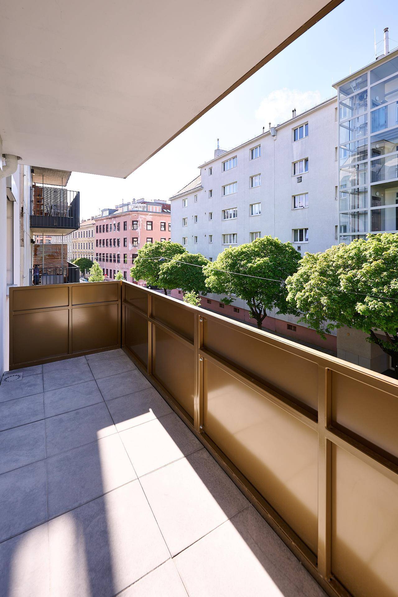 Sonniger Balkon mit Fliesenboden und Blick auf die umliegenden Gebäude und Bäume.