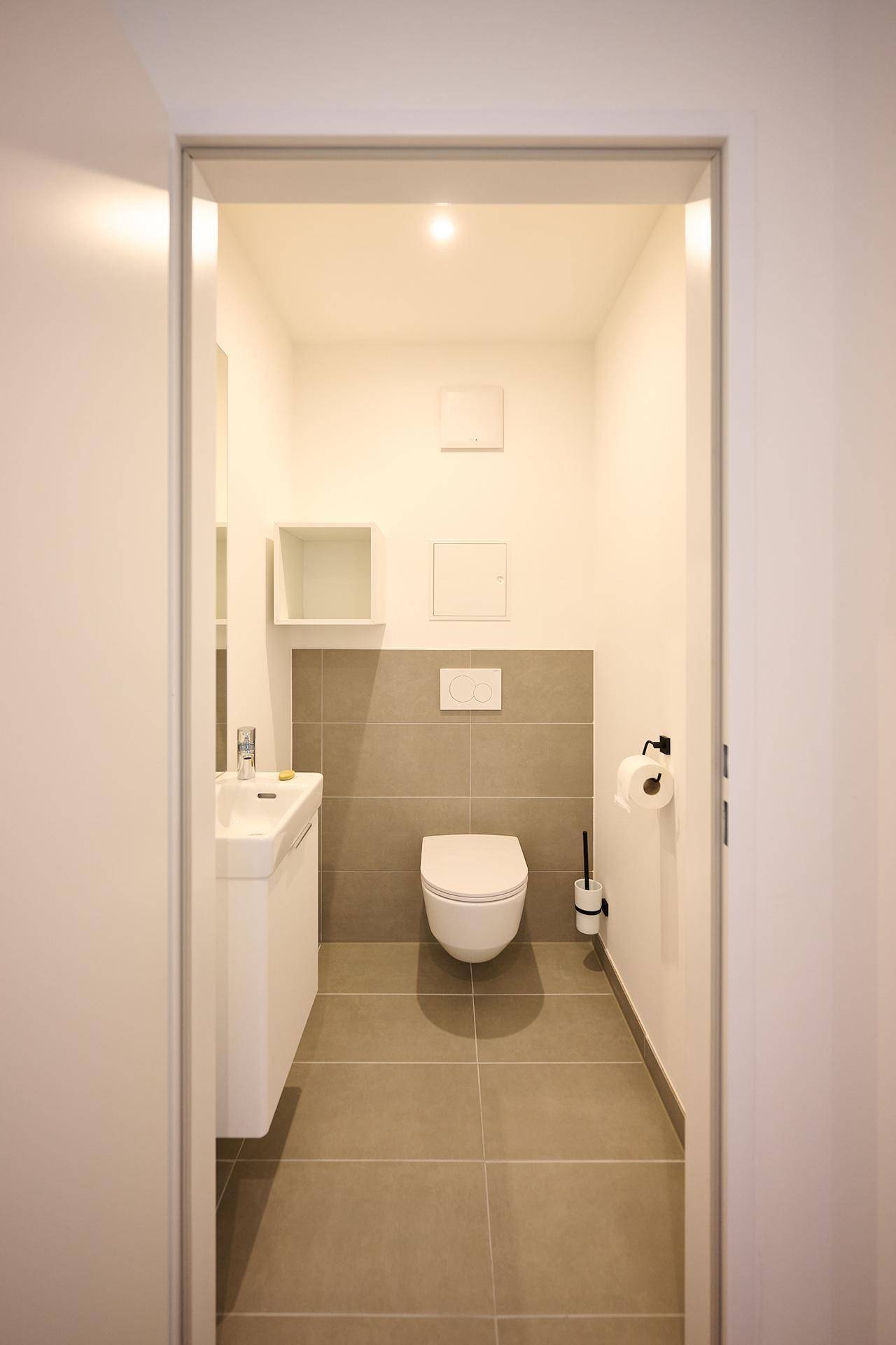 Modernes Gäste-WC mit Wand-WC, kleinem Waschbecken und grauen Fliesen.
