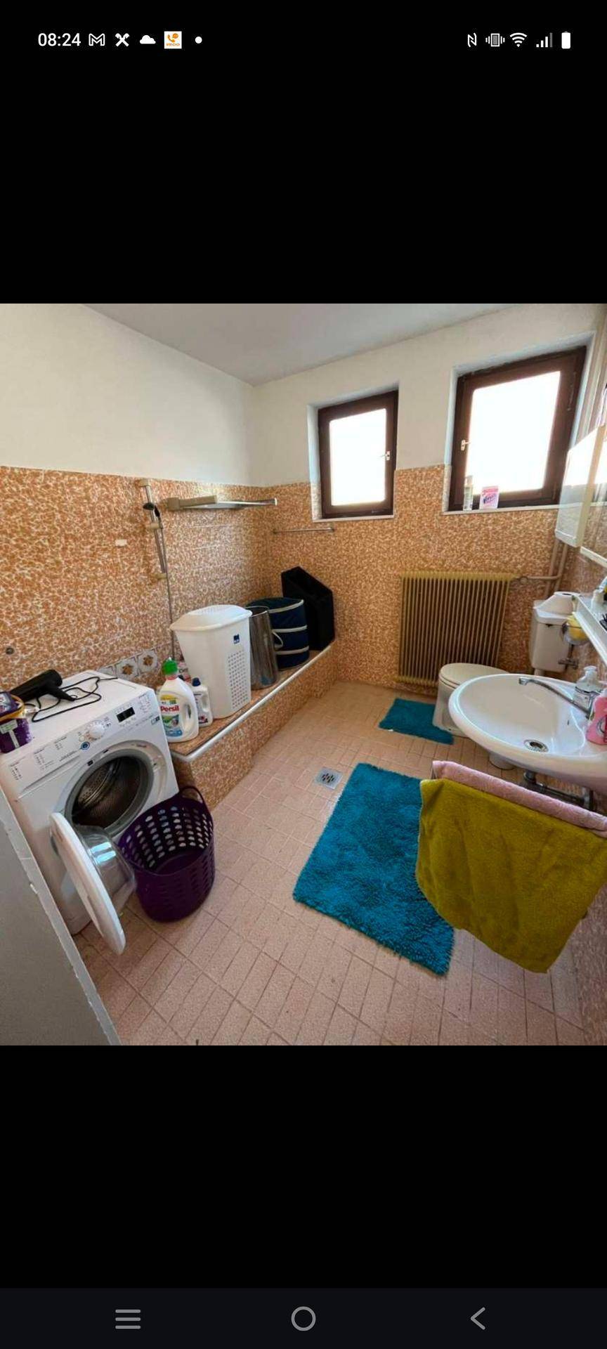 Badezimmer mit Waschmaschine, Waschbecken, Toilette und gefliesten Wänden und Boden in hellem Beige.