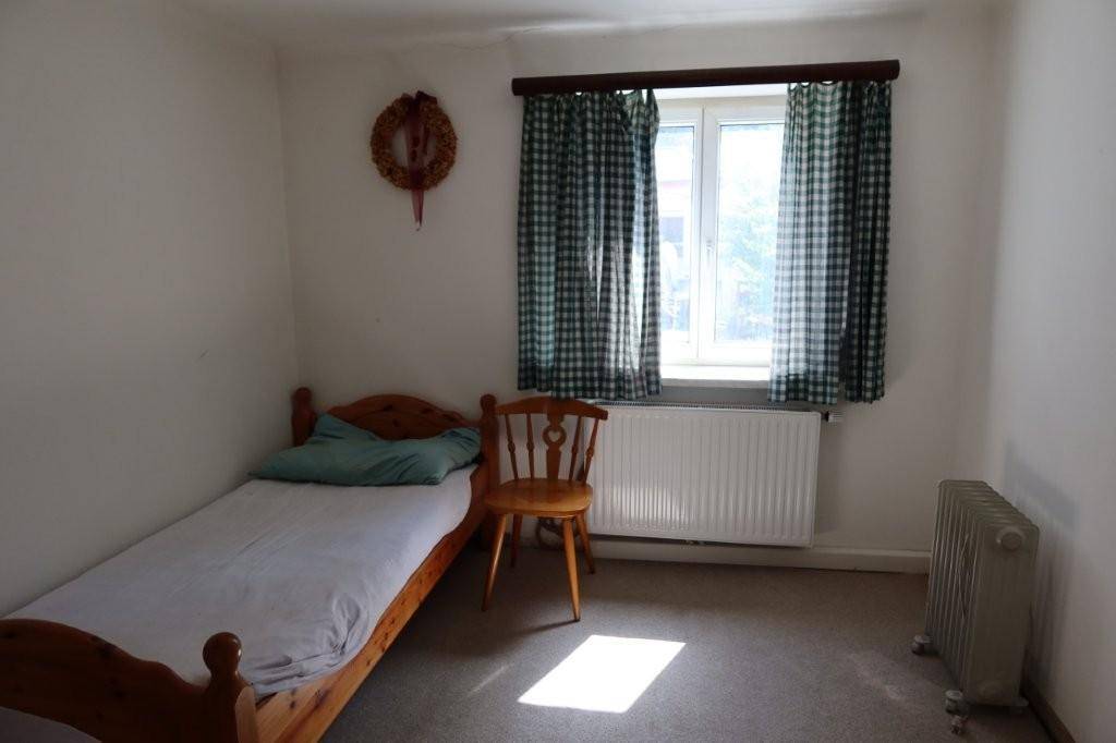 Einfaches Schlafzimmer mit Einzelbett, Teppichboden und Fenster mit karierten Vorhängen.