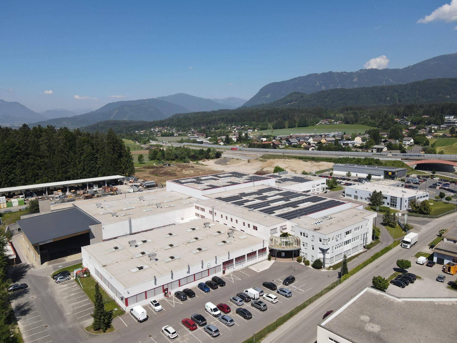 Großer Industriepark mit mehreren Gebäuden, Solaranlagen und einer beeindruckenden Bergkulisse im Hintergrund.