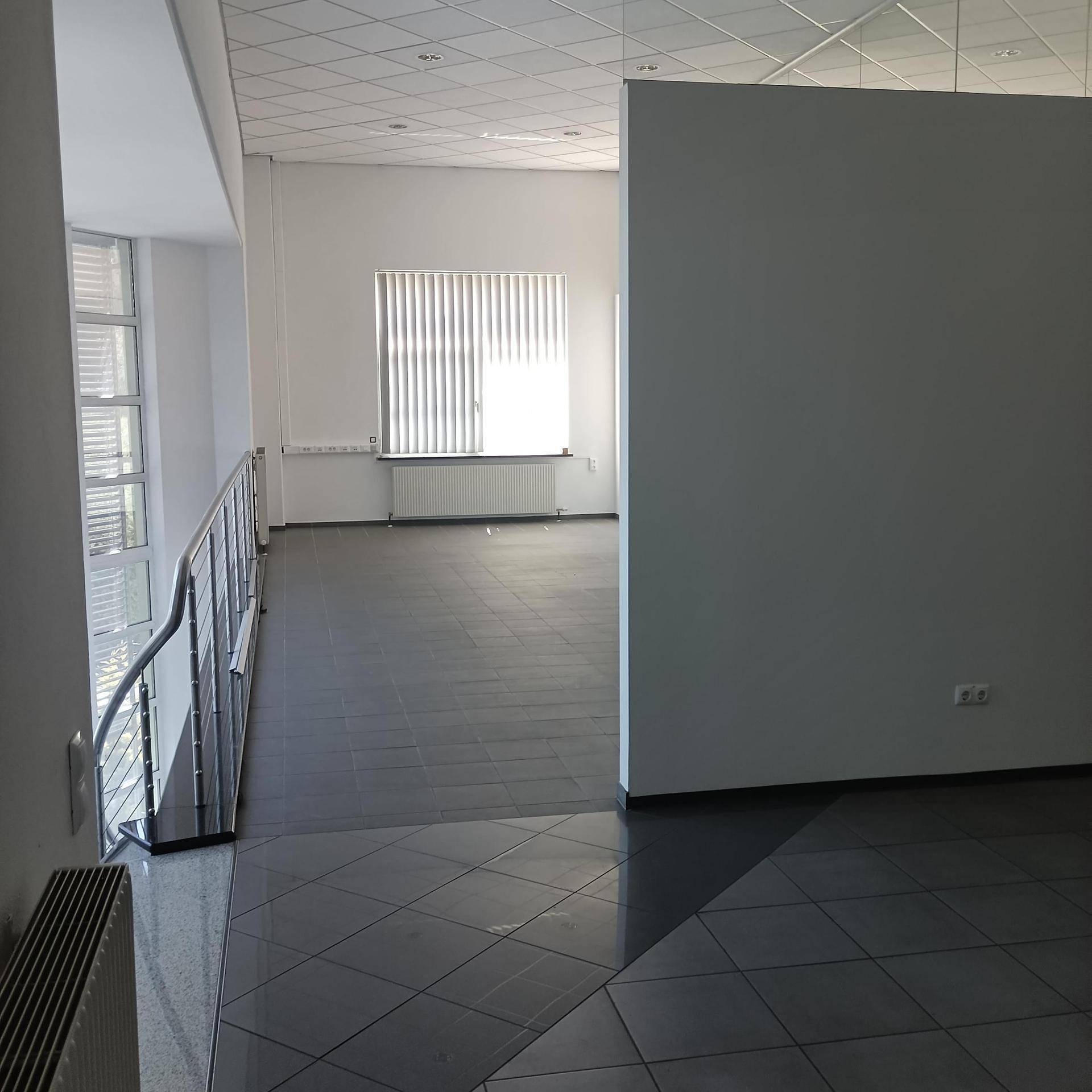 Heller Flur mit Fliesenboden und einem Geländer, das zu einem unteren Bereich führt, beleuchtet durch große Fenster.