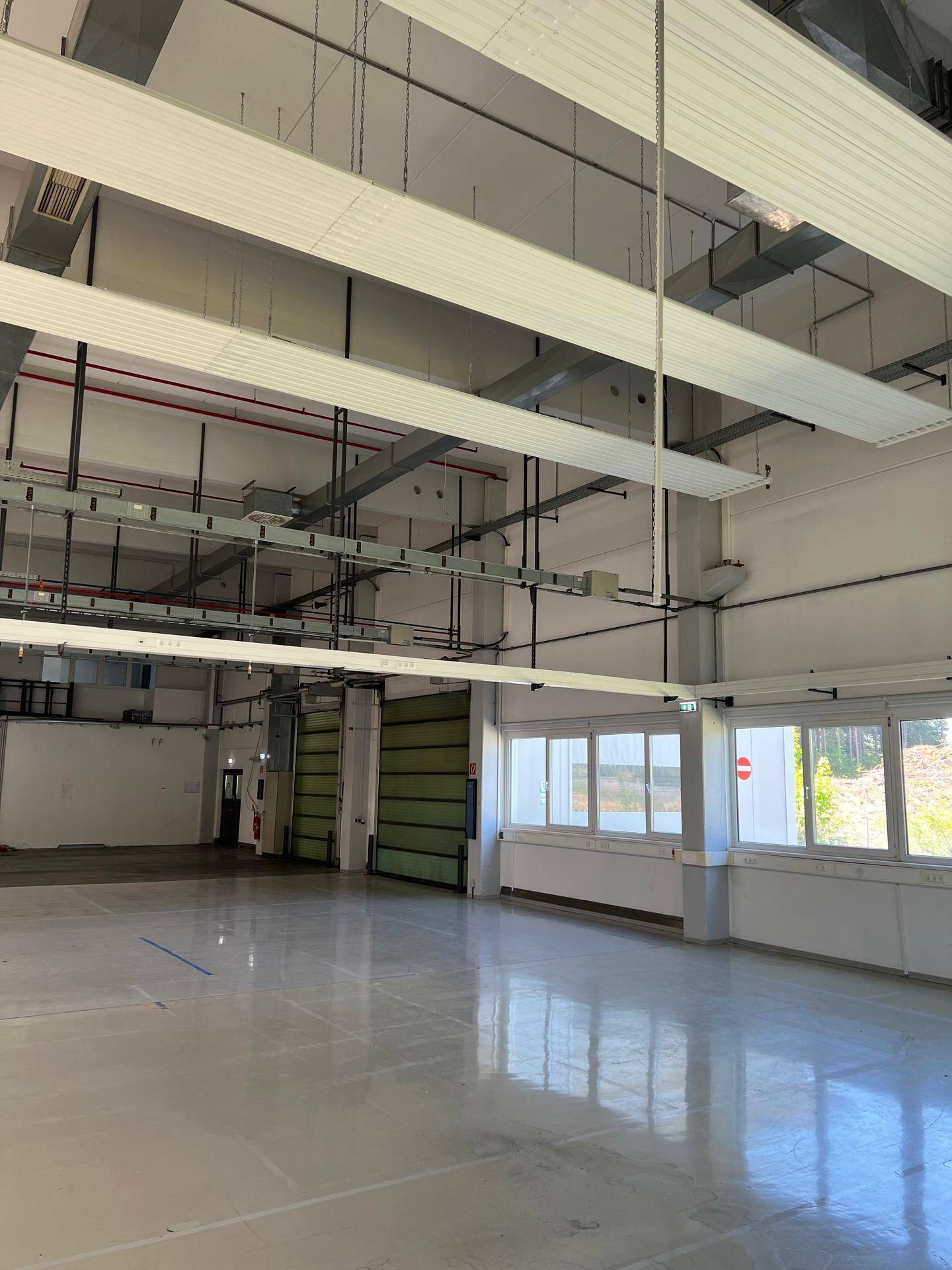 Geräumige Industriehalle mit großen Fenstern, Rolltor und sichtbaren Installationen an der Decke.