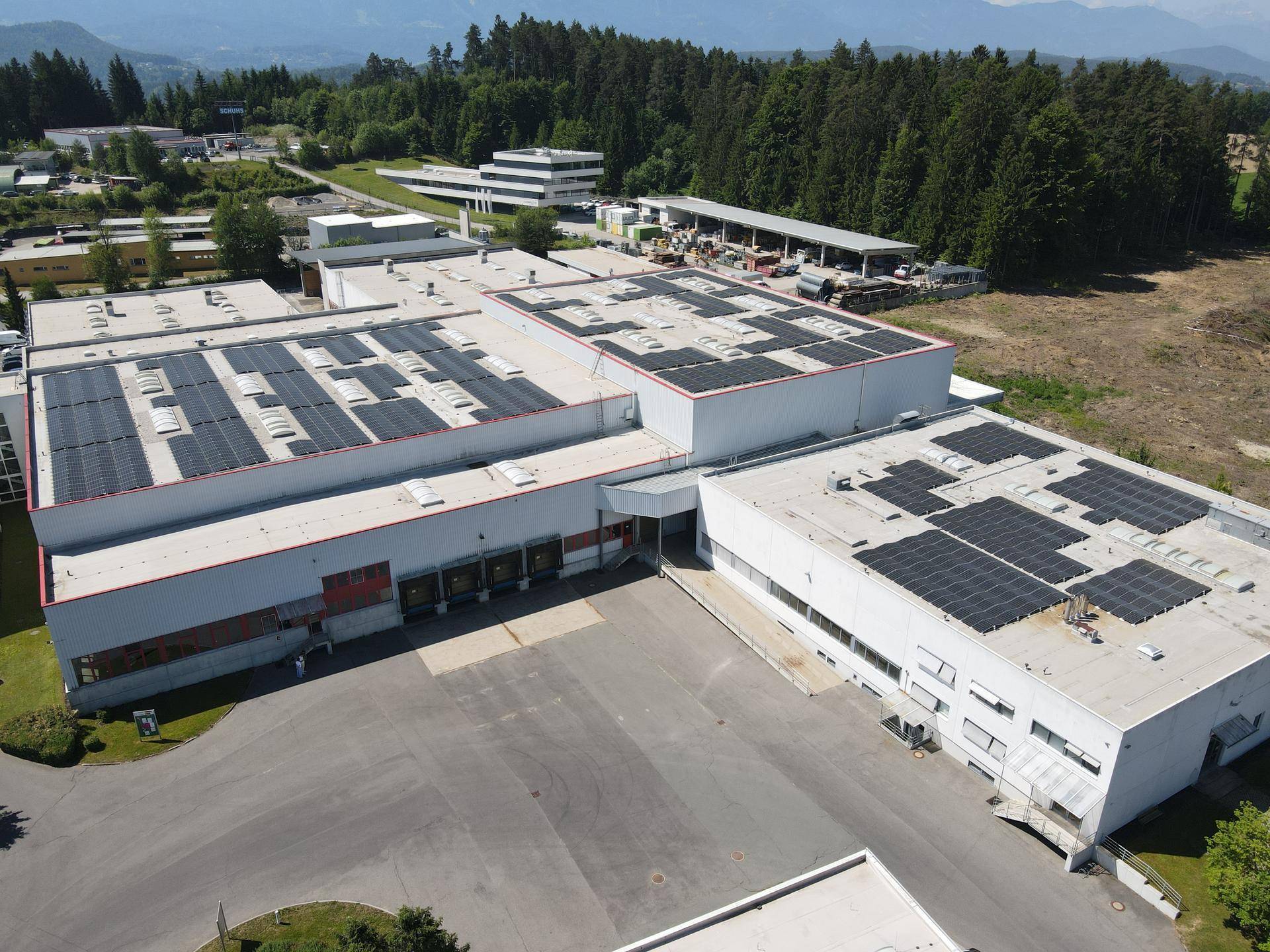 Luftaufnahme eines großen Industriegebäudes mit zahlreichen Solarmodulen auf den Dächern und umliegender Natur.