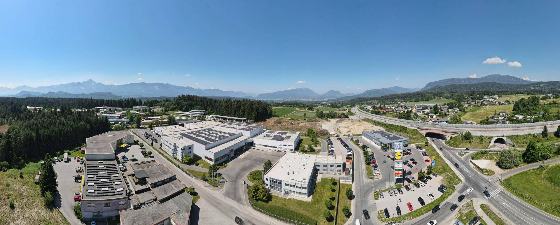 Panoramische Luftaufnahme eines Gewerbegebiets mit modernen Gebäuden, umgeben von Wäldern und Feldern, mit Autobahnanschluss und Bergblick im Hintergrund.