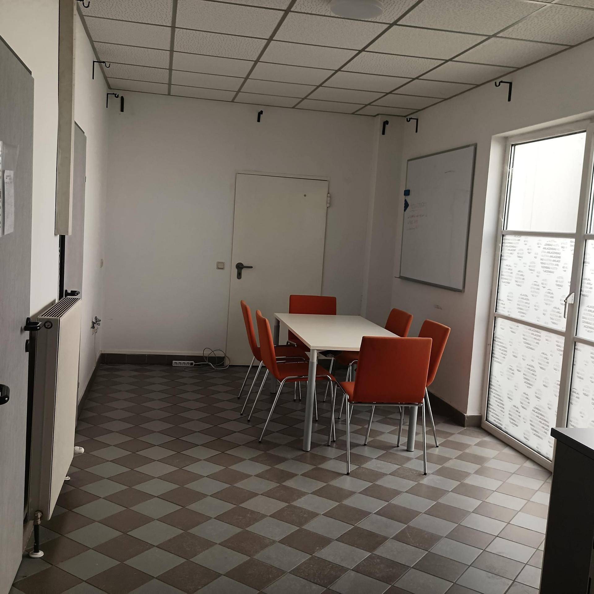 Essbereich mit kariertem Fliesenboden, einem weißen Tisch und orangefarbenen Stühlen, beleuchtet durch ein Fenster mit Jalousien.