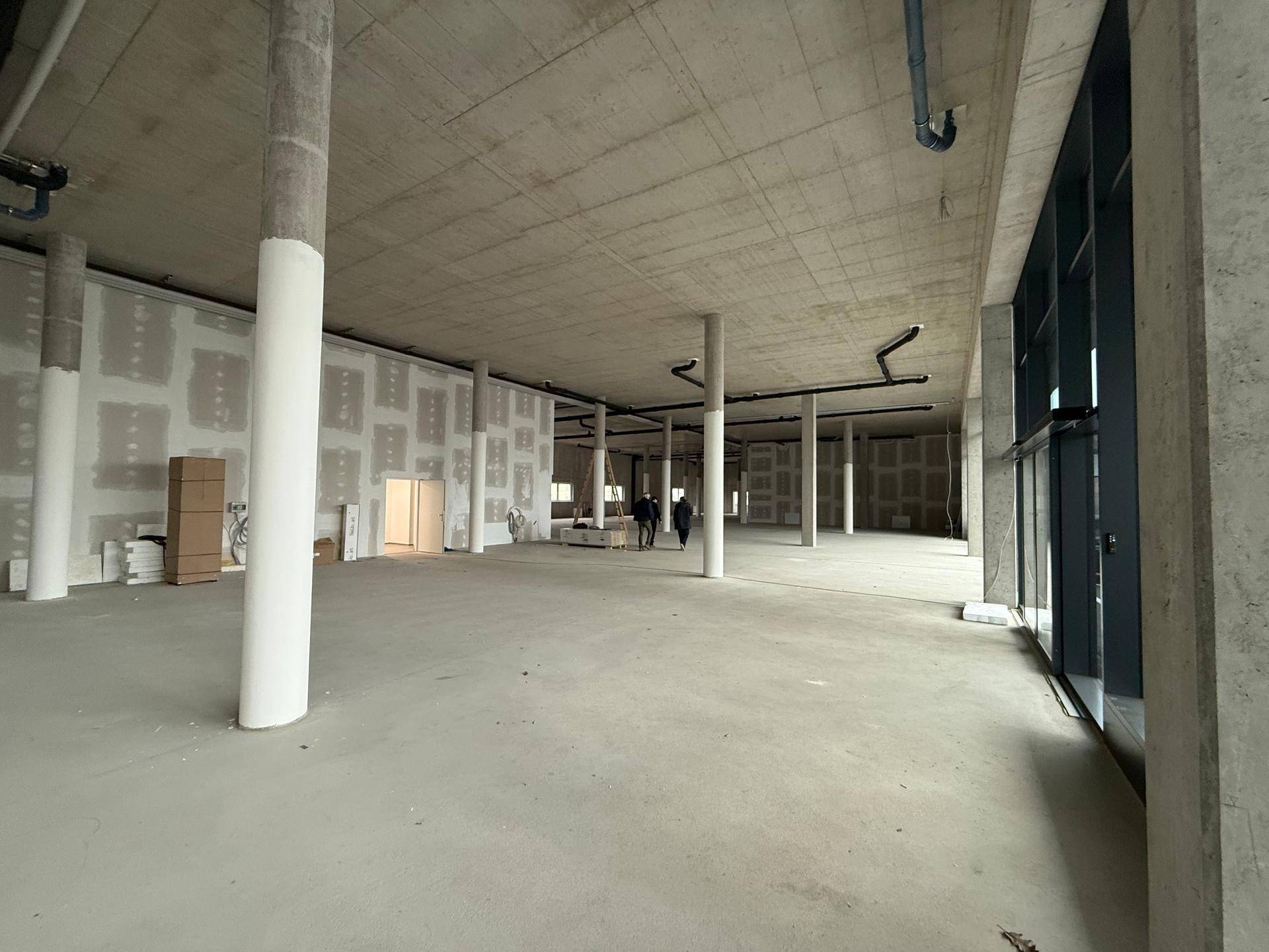 Weitläufiger, unfertiger Gewerberaum mit Betonsäulen, Trockenbauwänden und viel Tageslicht.