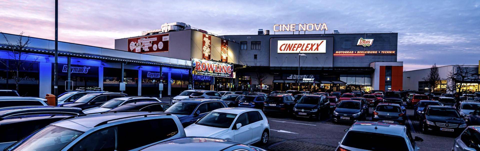 Abendliche Ansicht des Cine Nova und Cineplexx Gebäudes mit einem großen, belebten Parkplatz voller Autos.