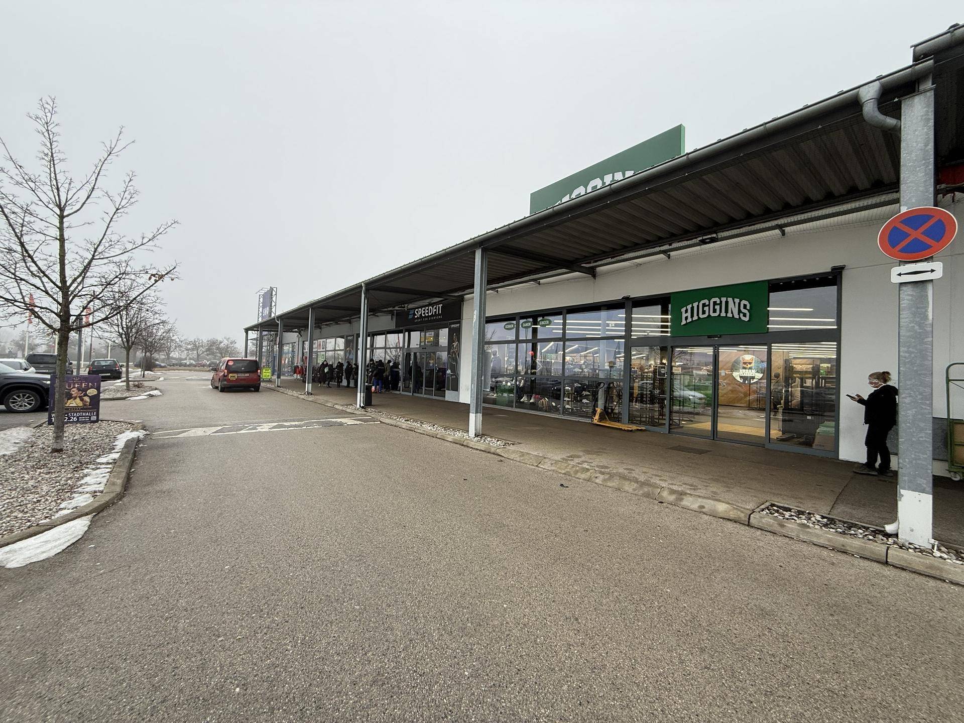 Lange Reihe von Geschäften in einem modernen Einkaufszentrum mit breitem Gehweg und Parkplätzen davor.