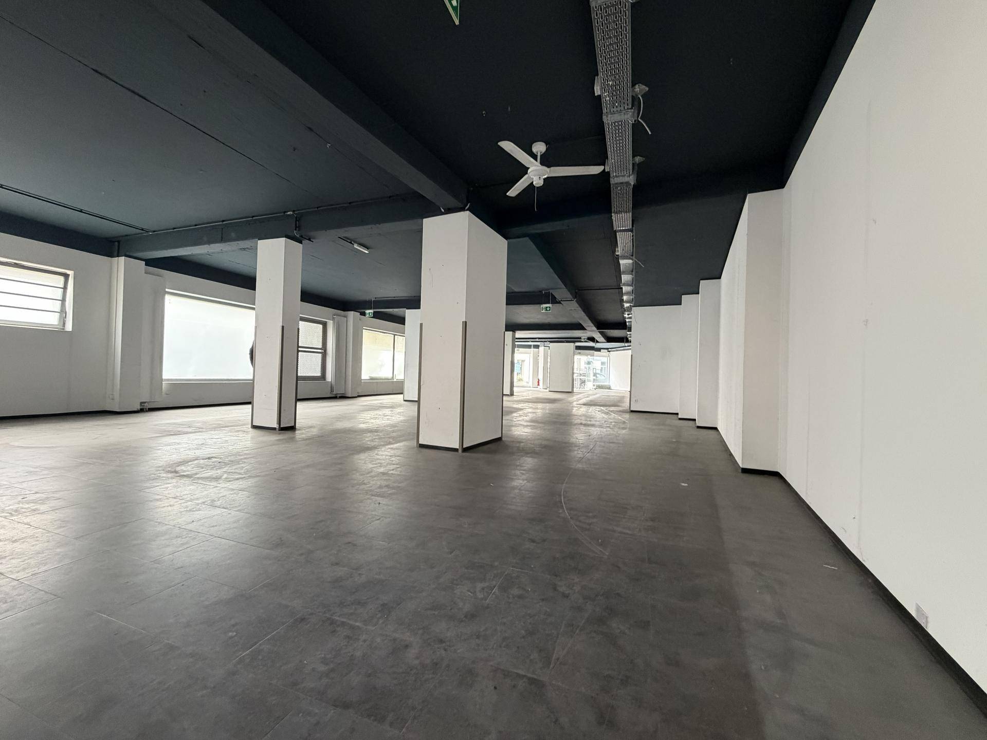 Geräumiger Geschäftsraum mit dunkler Decke, weißen Säulen und großen Fensterfronten, die viel Licht hereinlassen.