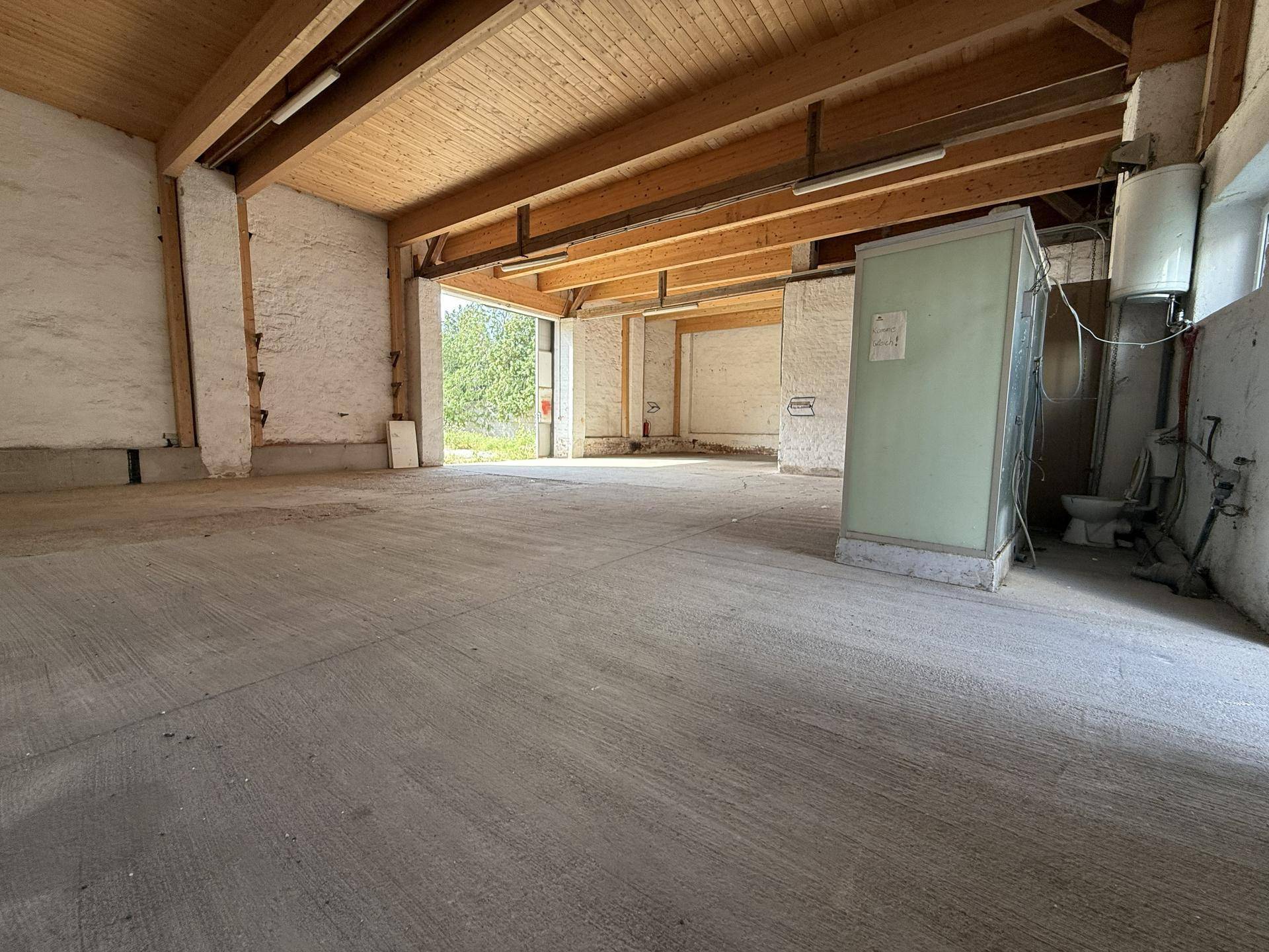 Großer, leerer Innenraum mit Betonboden, sichtbaren Holzträgern und weiß gestrichenen Ziegelwänden.