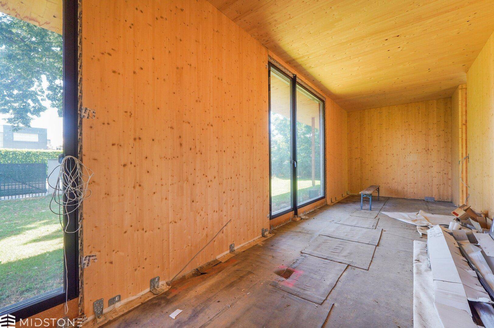 Innenraum im Rohbau mit Holzverkleidung, großen Fensterfronten und Blick auf die grüne Umgebung.