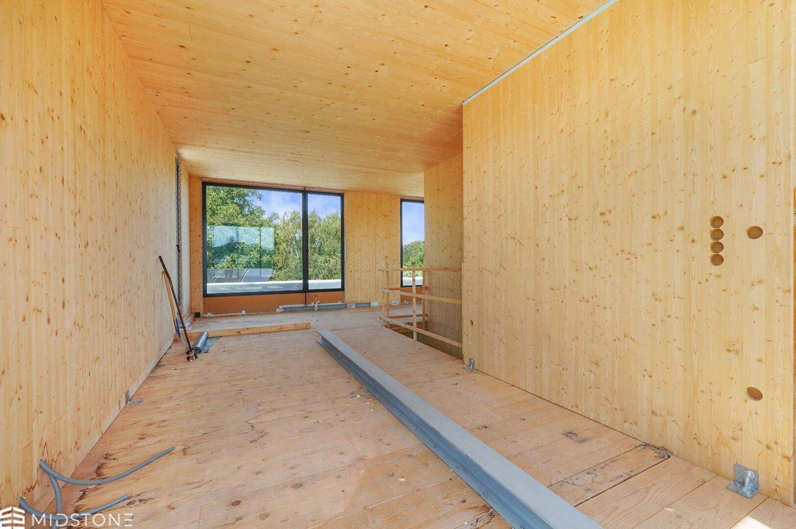 Geräumiger Innenraum im Rohbau mit Holzverkleidung und großen Fenstern, die viel Licht hereinlassen.
