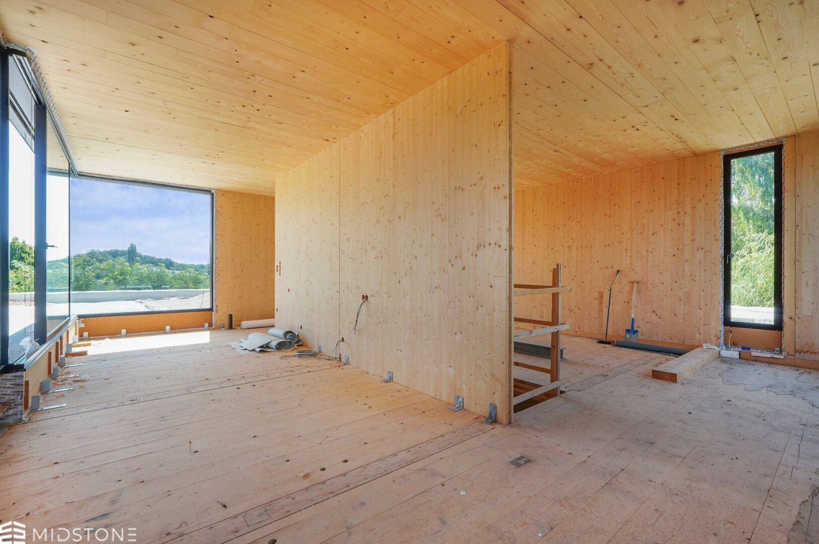 Rohbau eines Innenraums mit Holzverkleidung, großen Fenstern und Blick auf die grüne Umgebung.