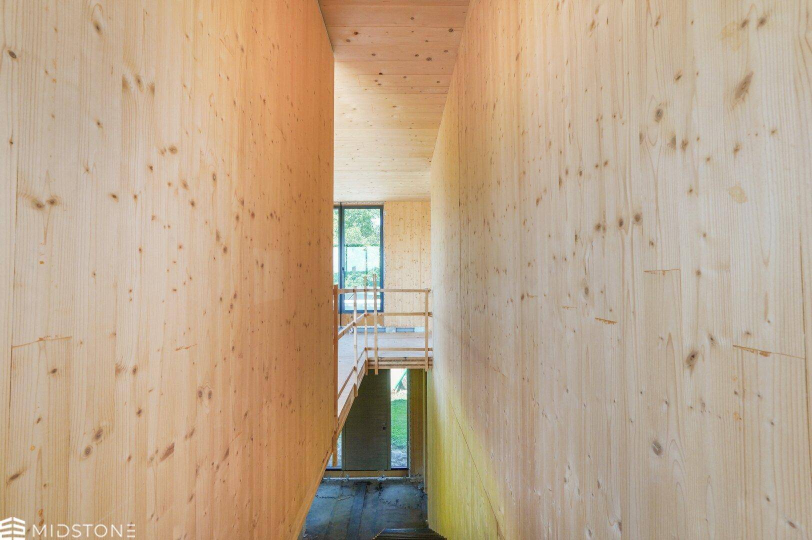 Schmaler Flur im Rohbau mit Holzverkleidung, der zu einer Treppe und weiteren Räumen führt.