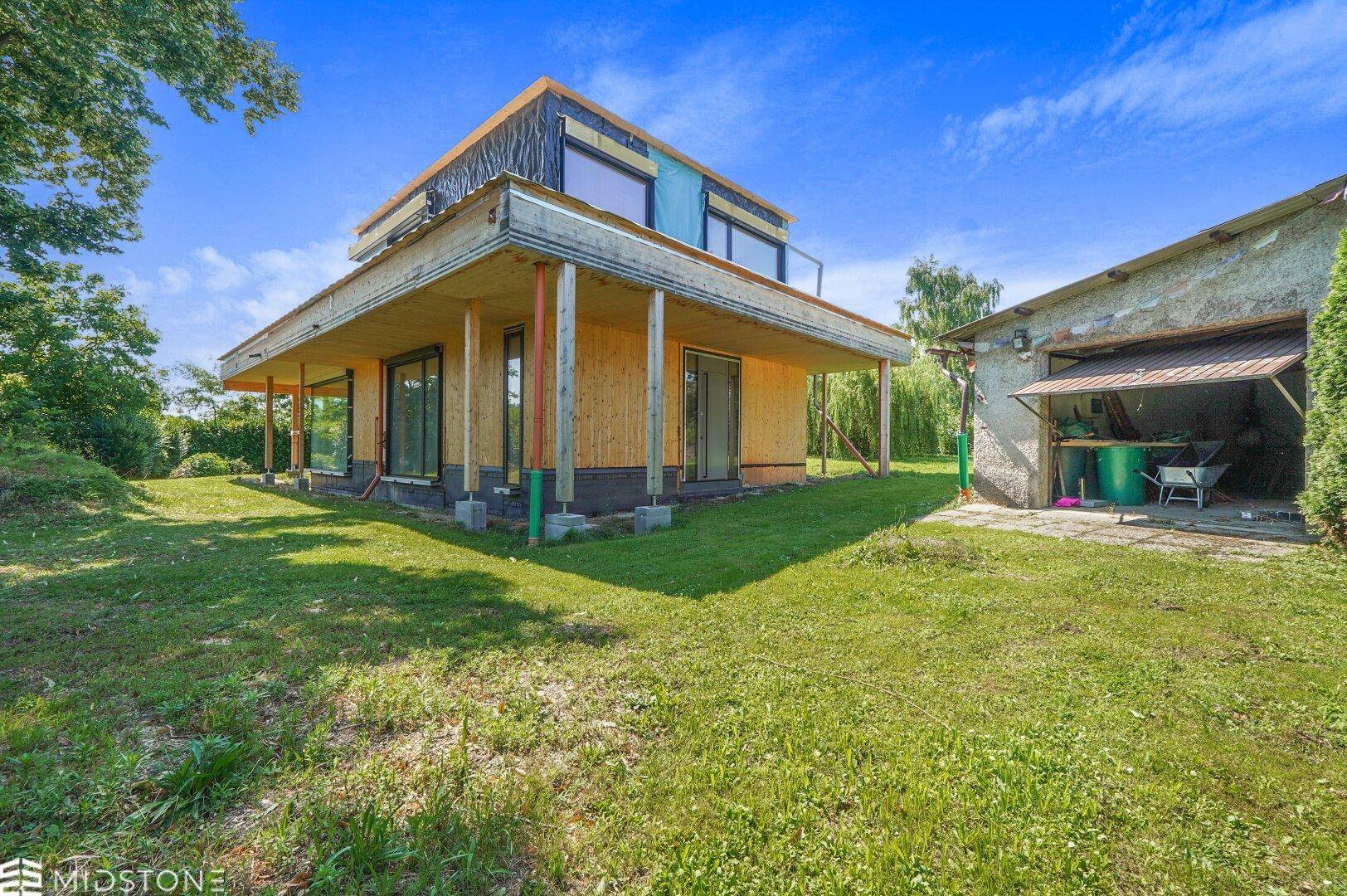 Neubauhaus in Holzbauweise mit großem Garten und angrenzendem Nebengebäude unter blauem Himmel.