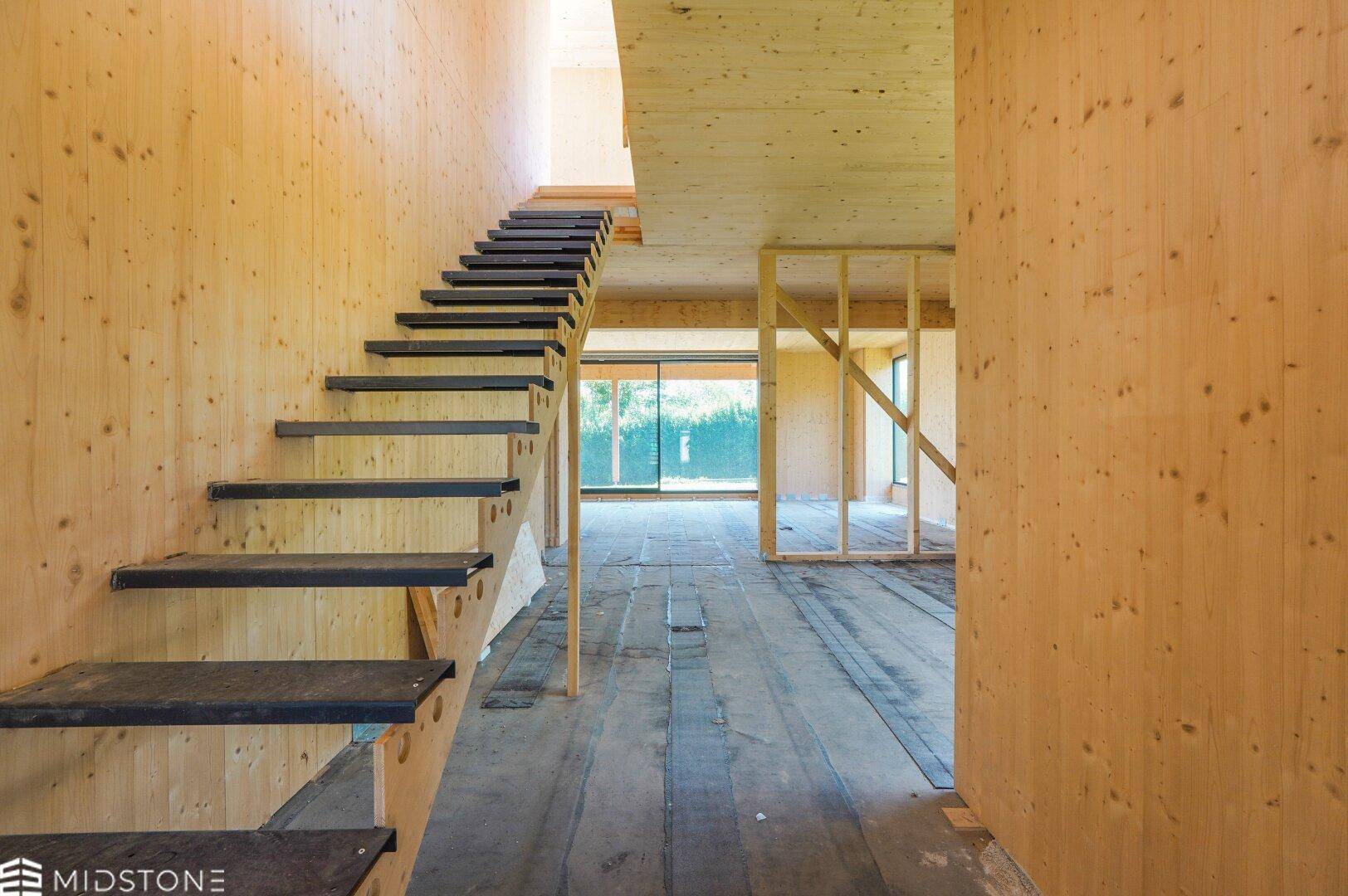 Innenraum im Rohbau mit Holzverkleidung und einer freitragenden Treppe, die in die obere Etage führt.
