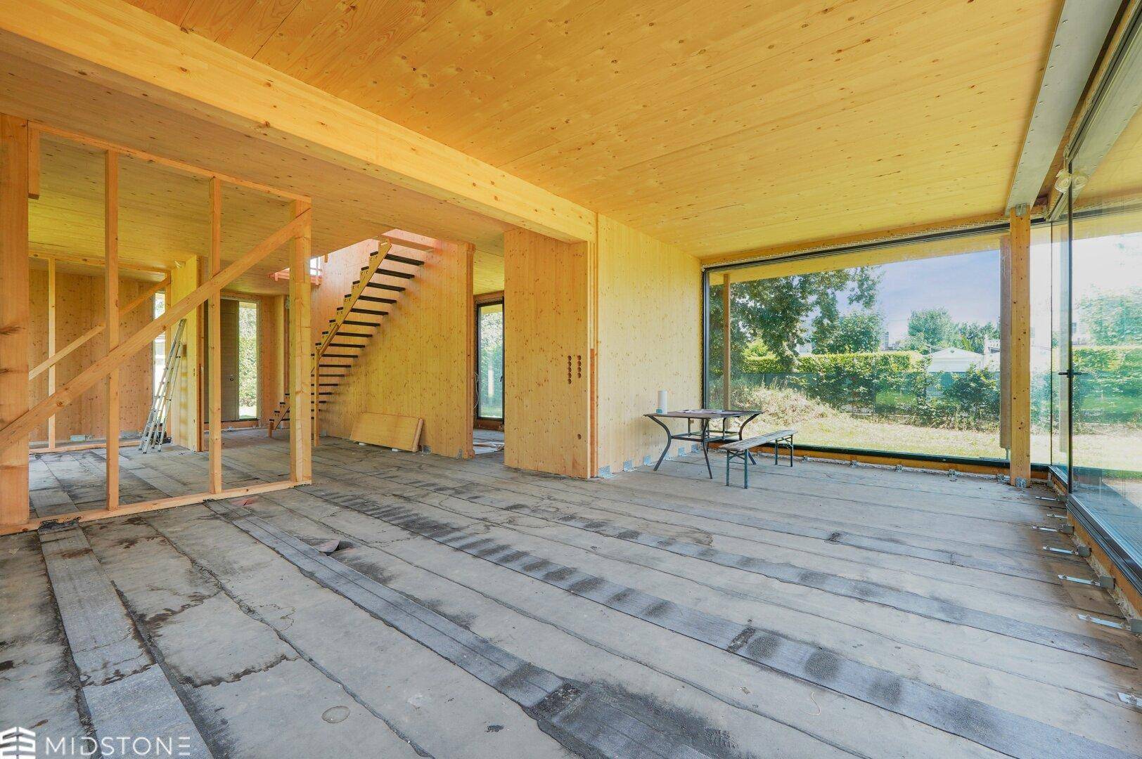 Geräumiger Innenraum im Rohbau mit Holzverkleidung, großen Fenstern und sichtbarer Treppenkonstruktion.